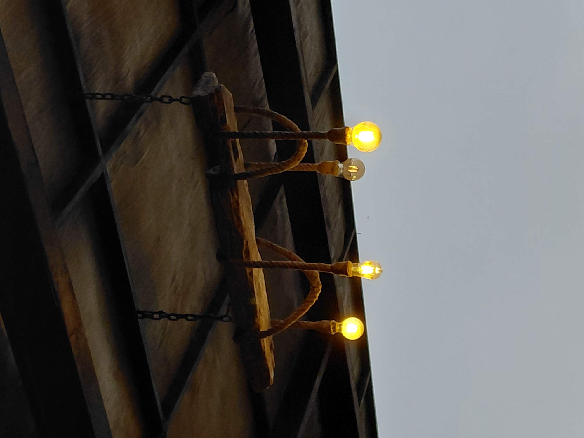 A rustic chandelier made of a wooden plank and thick ropes hangs from a dark, tiled ceiling in Kintamani, Indonesia. Four Edison-style light bulbs, emitting a warm yellow glow, are suspended from the ropes. The ceiling appears to be part of an outdoor or semi-outdoor structure, possibly a patio or pergola, given the bright, overcast sky visible below the chandelier. The overall impression is one of a casual, rustic ambiance.