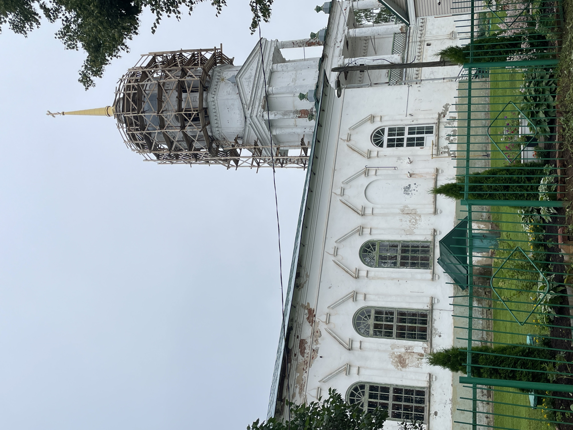The image shows a white Orthodox church with a golden cross on top, situated in Kstinino, Russia. The church's dome is partially covered in scaffolding, suggesting it is undergoing renovation. The church has a long, white facade with arched windows that have green frames. A green metal fence encloses the front of the church, with manicured bushes and flowers visible behind it. The sky is overcast, and a few trees can be seen on the right side of the image. There are no people visible in the image.