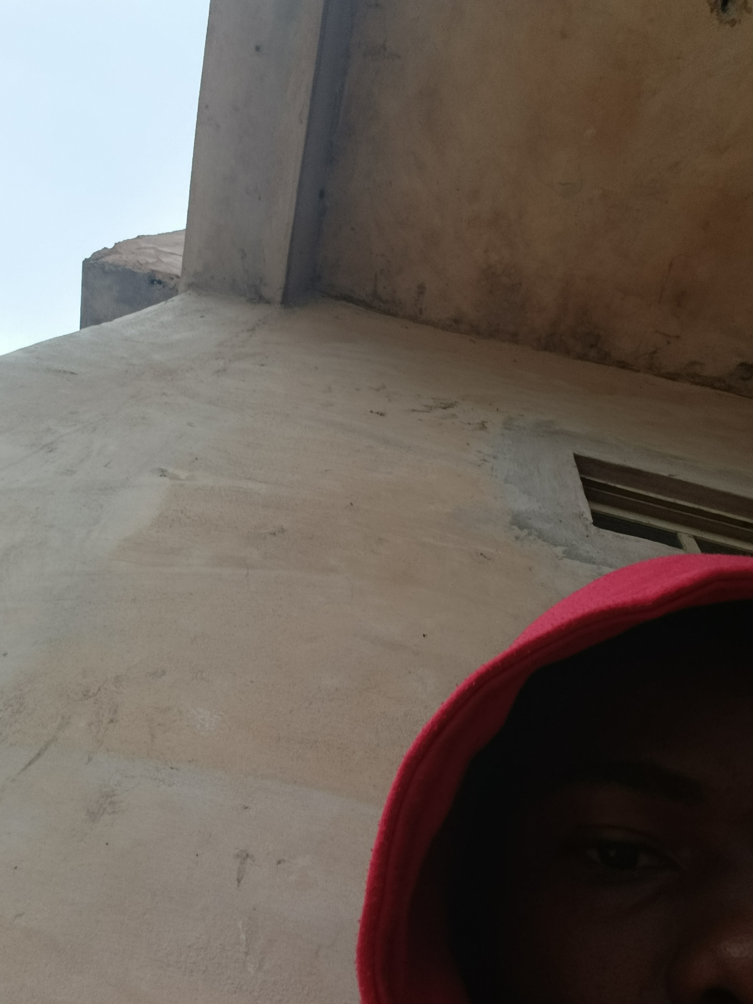 The image is a close-up, low-angle shot taken in Ukpor, Nigeria. It features the upper part of a person's face, partially obscured by a red hooded garment, suggesting a selfie. The background consists of a rough, textured concrete wall and a section of a lighter colored, possibly stuccoed wall with a small, dark rectangular window opening. Above the walls, the sky appears pale and overcast. The lighting is subdued, casting the lower part of the face in shadow and highlighting the texture of the building materials.