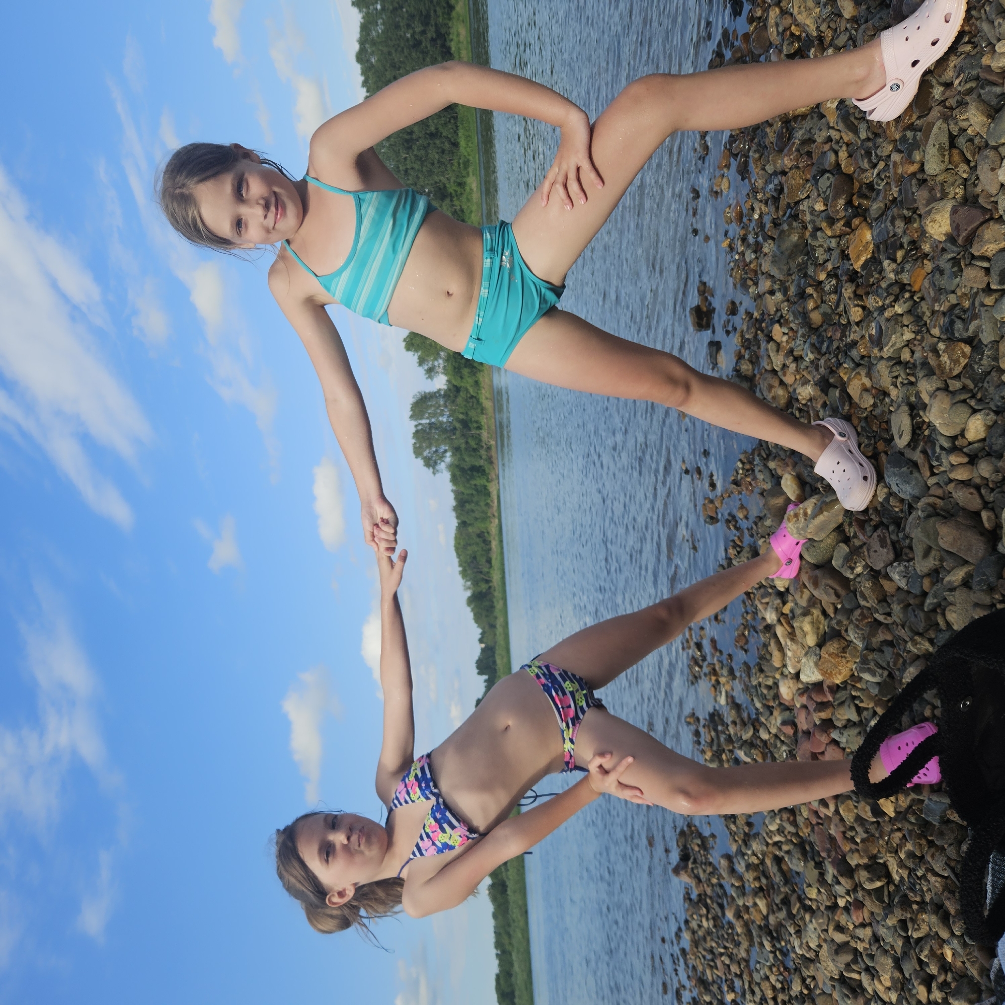 Two young girls are posing on a rocky shore by a body of water, likely a river, given the lush green trees lining the opposite bank. The sky above is a bright blue with scattered white clouds, suggesting a sunny day, possibly late afternoon or early evening due to the lighting.
The girl in the foreground, wearing a patterned bikini, is reaching out to hold hands with the other girl. She has her legs spread and is looking towards the camera with a playful expression. The second girl, dressed in a striped turquoise bikini, is standing with one leg extended and her hand on her hip, also looking at the camera. Both girls are wearing colorful clogs on their feet, one pink and one light pink.
The ground is covered in a variety of smooth, multi-colored stones, and the water appears calm. The overall scene depicts a relaxed and fun moment during what seems to be a summer outing by the water. There is no visible text in the image.