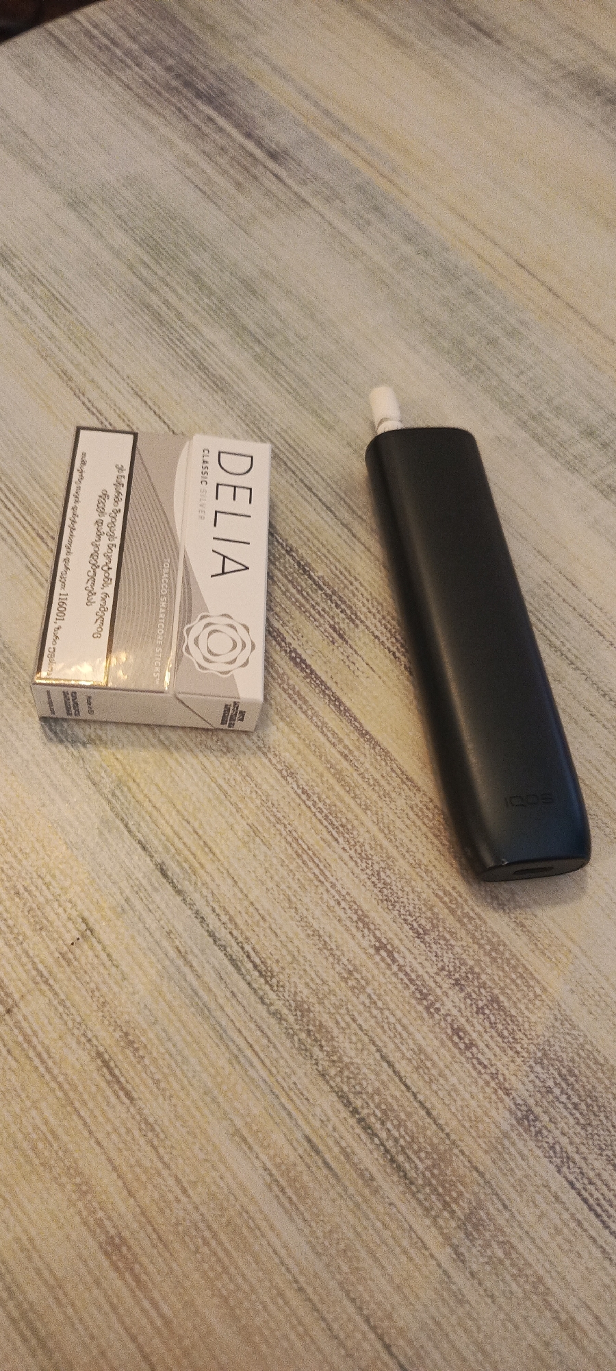 This is a close-up, eye-level, full-shot photograph of a black IQOS device and a pack of "DELIA CLASSIC SILVER" tobacco sticks placed on a wooden surface. The wooden surface has a light brown and grey striped pattern. The IQOS device is positioned to the right of the pack of tobacco sticks and is mostly in focus, with the IQOS logo visible on its side. The pack of tobacco sticks is to the left, slightly out of focus. The pack is white and grey with text and a circular floral design. Some text on the pack appears to be in Georgian script. The image appears to have been taken indoors, possibly in an apartment in Rustavi, Georgia, though this cannot be definitively confirmed by the image itself. There are no people or animals present in the image. The lighting suggests it was taken in a well-lit room.