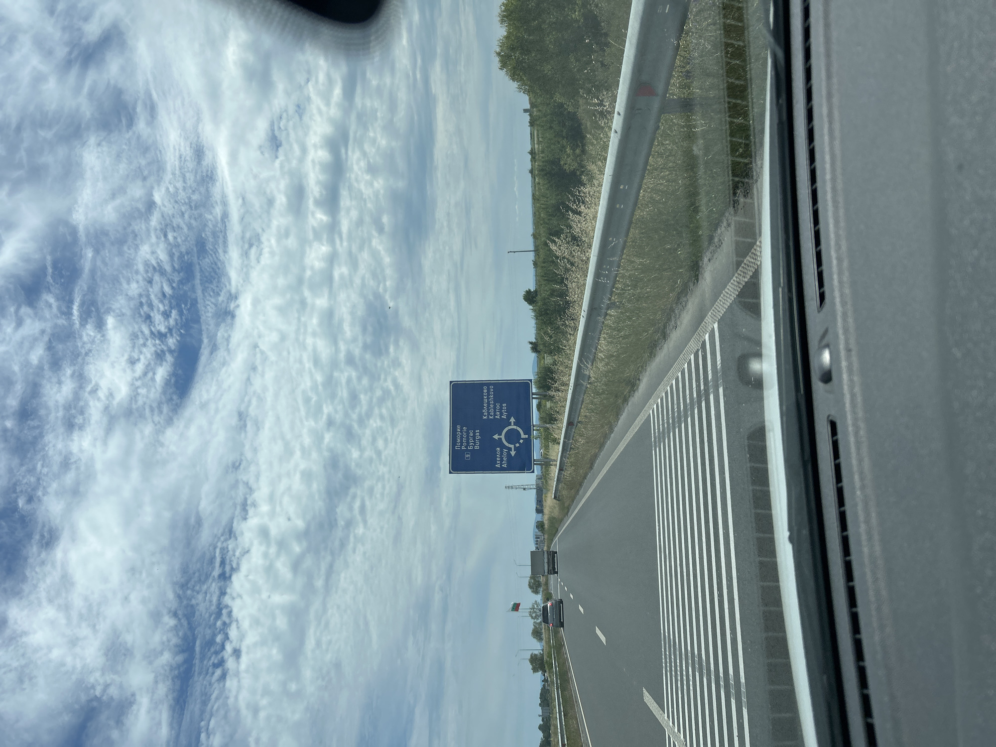 The image depicts a view from inside a vehicle, looking forward along a highway. The road is paved with multiple lanes, marked by white lines. A highway sign, blue with white lettering and a circular arrow diagram, is visible on the right side of the road. The sign indicates directions to "Pomorie," "Burgas," "Kableshkovo," and "Aheloy." The landscape beside the highway consists of dry grass and scattered trees under a cloudy sky. Two cars and a truck are on the highway ahead. A Bulgarian flag is planted on a pole to the left of the road.