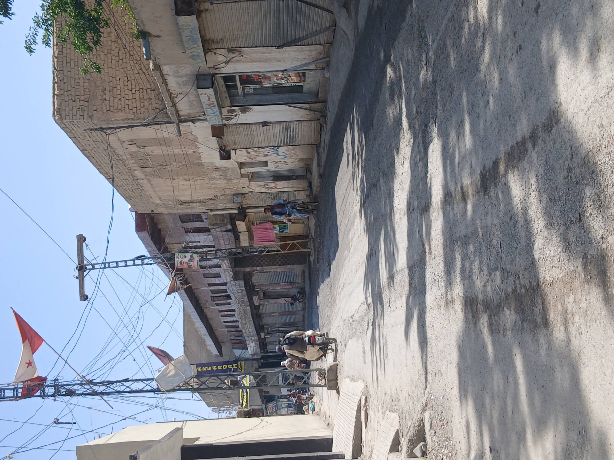 The image depicts a street scene in Quetta City Tehsil, Pakistan. The street is lined with buildings, many of which have closed metal shutters indicating closed businesses. Wires and electrical poles are visible overhead. People are riding motorbikes on the street. Flags are hanging from the poles. A shadow of trees is on the ground.