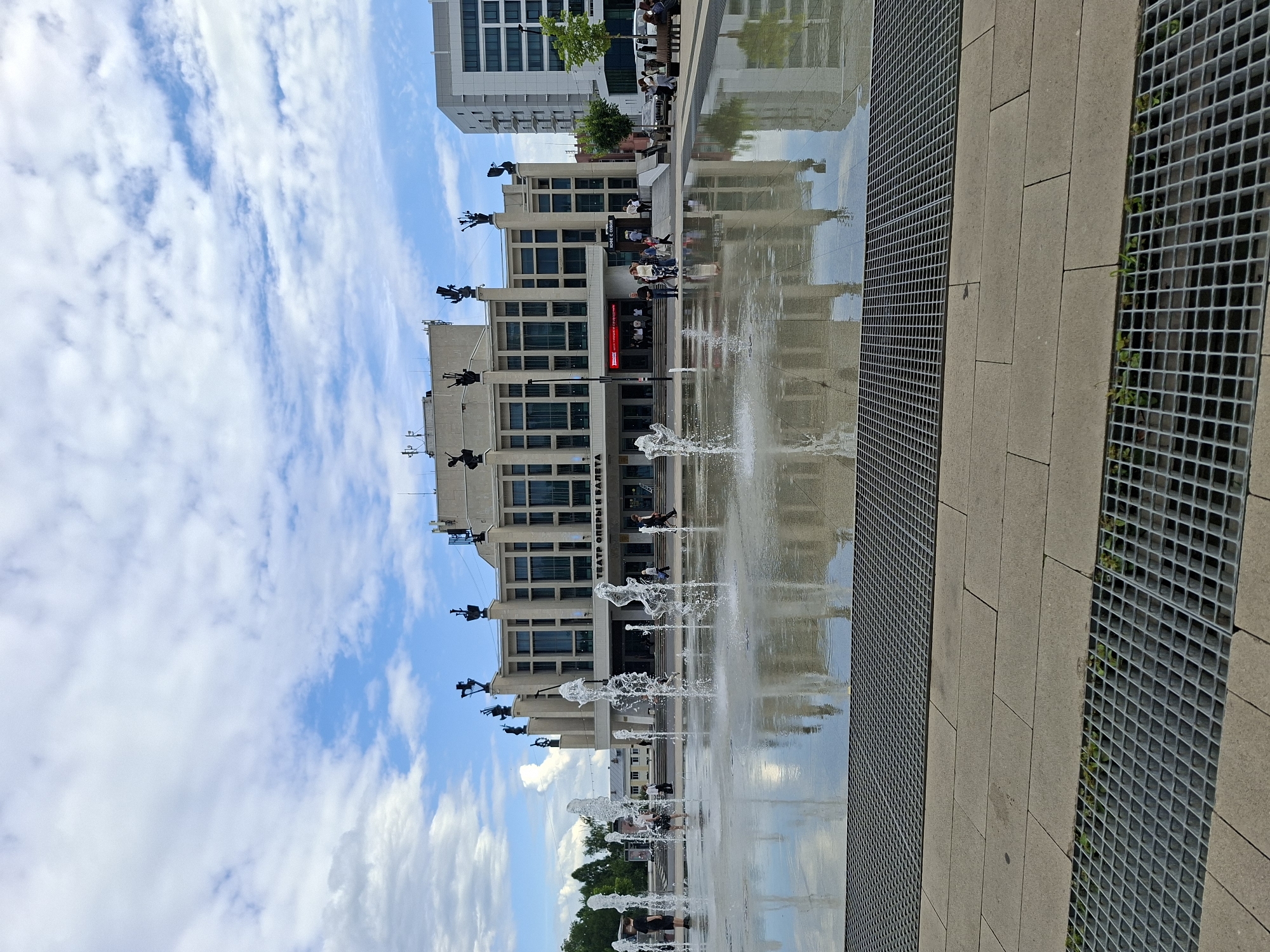 This media file captures an outdoor scene in Izhevsk, Russia, featuring a modern building with architectural sculptures adorning its facade and a fountain in the foreground. The building, likely a public space, is characterized by a repeating pattern of windows and several prominent sculptures depicting human figures in dynamic poses. Water jets from a fountain create a playful and refreshing atmosphere, with their reflections shimmering on the wet pavement.
Several people are visible in the scene, some interacting with the fountain, perhaps enjoying the cool water on a warm day, while others are seen walking or sitting, contributing to the sense of urban life. The weather appears to be pleasant, with a bright blue sky dotted with white, fluffy clouds, suggesting a daytime setting. The foreground includes a paved area with metal grates, likely part of the fountain's drainage system, and stone tiling.
Notable text visible on the building reads "ТЕАТР ОПЕРЫ И БАЛЕТА," which translates to "Theater of Opera and Ballet." This text clearly identifies the building's purpose and provides a significant location cue. The overall mood conveyed by the image is one of leisure and urban vibrancy, with the architecture and the lively fountain creating an engaging public space. The reflection of the building and the sky on the wet ground adds a layer of visual interest and depth to the scene.