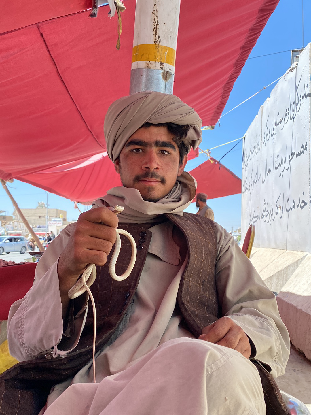 A man is sitting under a red awning in the city of Kandahar, Afghanistan, holding a small, white snake in his right hand. He is wearing a tan turban, a light gray shirt, and a brown vest. A white pole with a yellow stripe is positioned above him. Behind the man is a white wall with Arabic writing on it, and a blue sky is visible above. In the background, there are cars and other people, suggesting an urban setting.