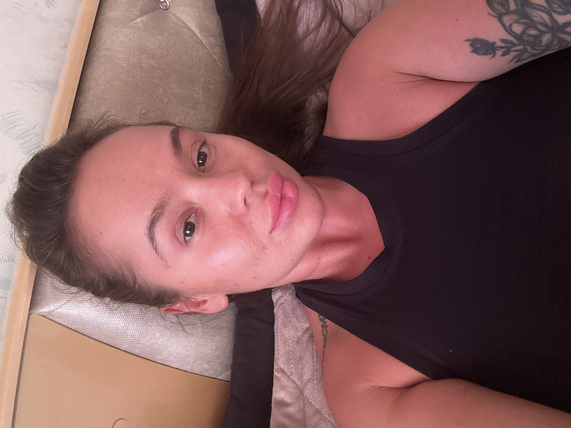 A young woman is lying on a bed, with her head resting on a pillow. She is wearing a black tank top. Her hair is dark brown and pulled back. She has full lips and dark eyes. There are tattoos on her left shoulder and arm. The bed has a light brown headboard and a textured gray duvet. The overall lighting suggests it is daytime, with a warm, rosy hue.