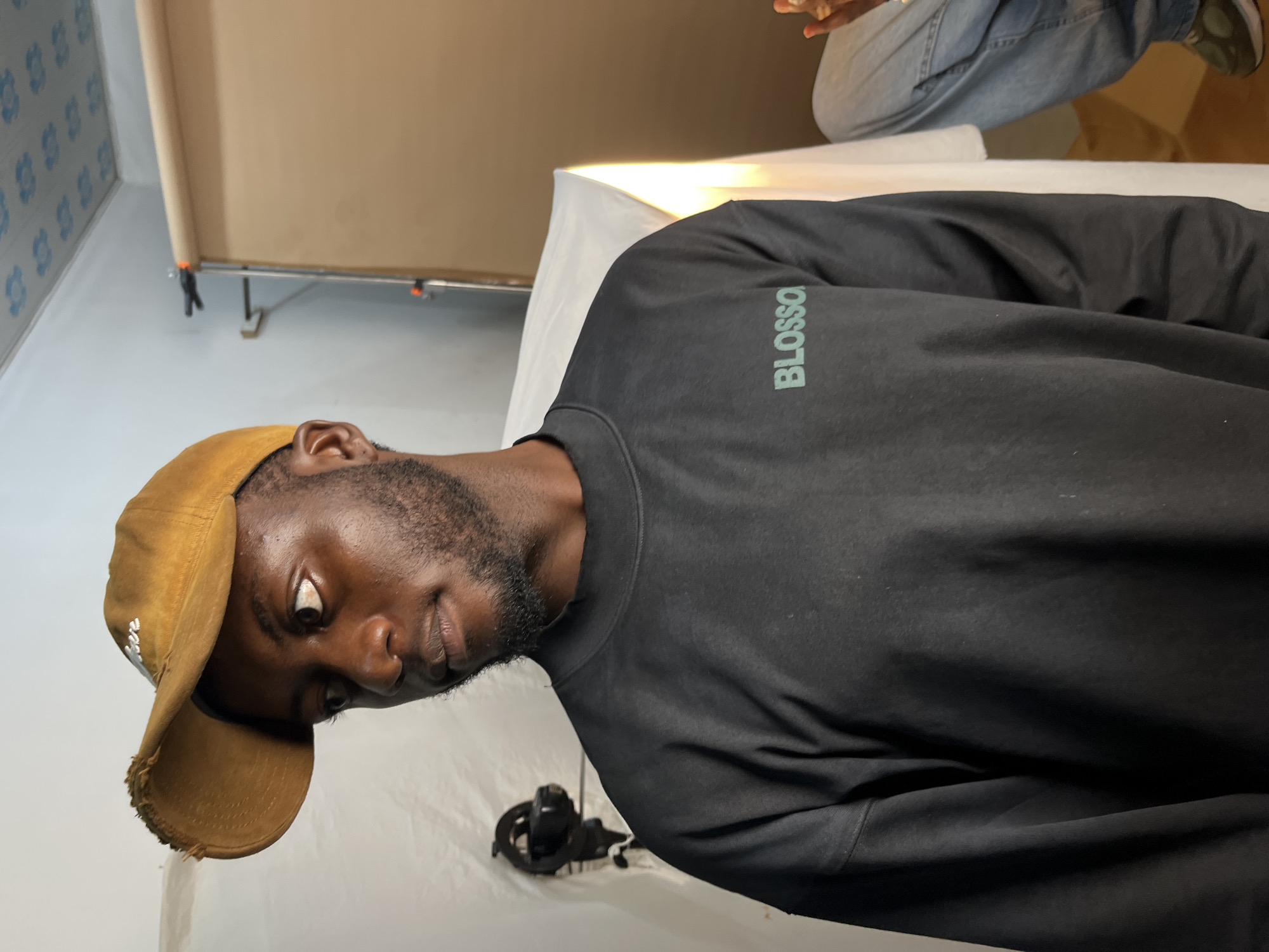 A medium shot captures a Black male wearing a mustard yellow baseball cap and a black long-sleeved shirt with "BLOSSO" printed in light green on the chest. The man is looking to his left with a neutral expression. He has a full beard and short, dark hair. In the background, a white backdrop is set up with a light brown roll of paper behind it, suggesting a studio environment. To the left, a black studio light is visible. Further to the right, another person is partially visible from the waist down, wearing blue jeans and green sneakers. The ceiling is decorated with blue and white floral tiles. The scene appears to be an indoor studio, likely in Gura Top, Nigeria.