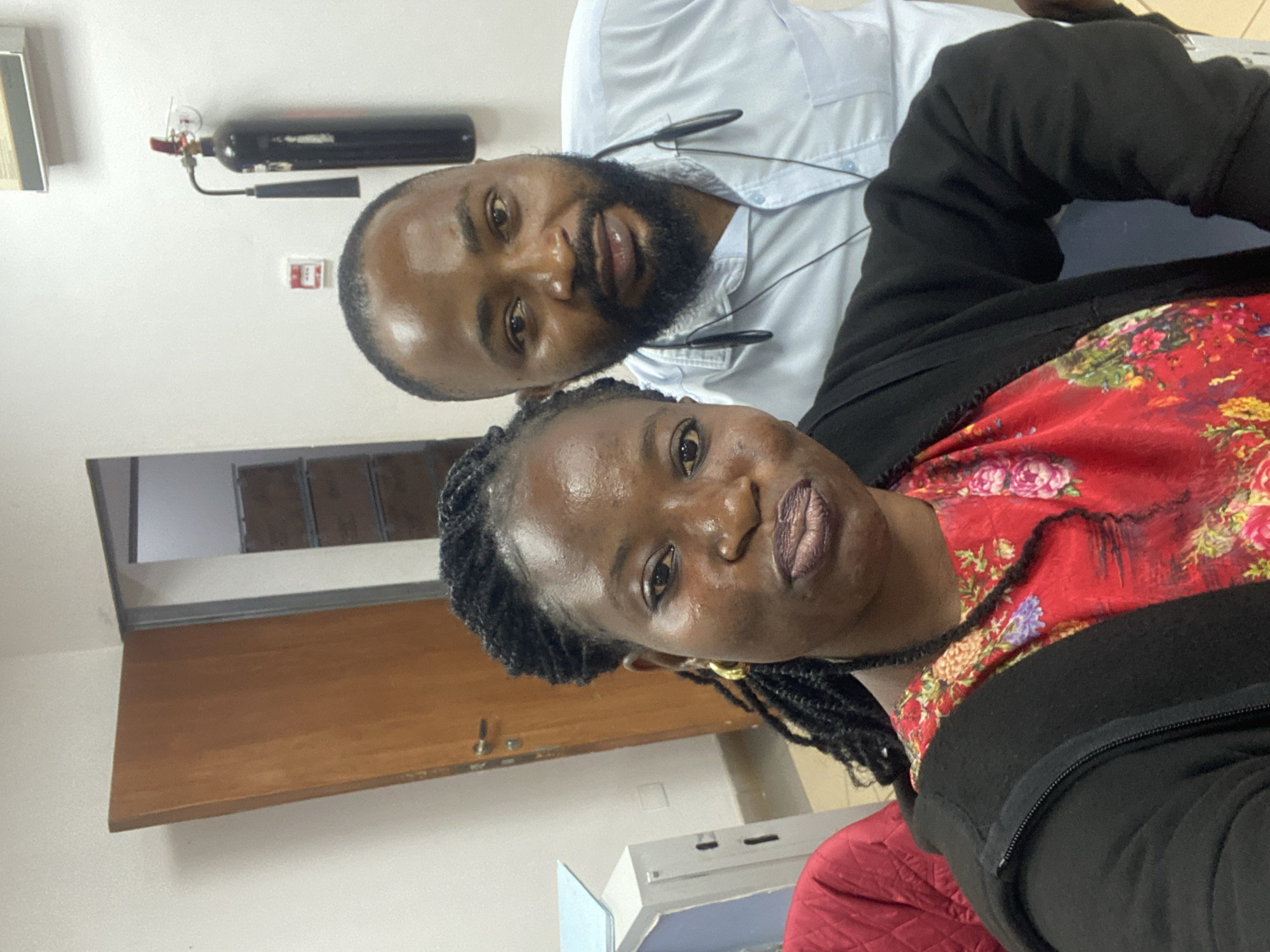 An indoor, medium shot captures a selfie of two people, a woman and a man, in what appears to be an office or storage area. The woman is in the foreground on the left, her face angled slightly towards the camera. She has dark skin, braided hair that falls over her shoulders, and is wearing large gold hoop earrings and a red dress with a floral pattern. She has on a black zip-up hoodie over the dress. Her lips are pursed. The man is positioned behind her and to her right, his face also angled toward the camera. He has dark skin, a beard and mustache, and is wearing a light blue collared shirt with earbuds around his neck. In the background, a wooden door is visible on the left, and to the right, a black fire extinguisher is mounted on the wall. Further in the background, there appear to be shelves with items. The lighting is bright and even.