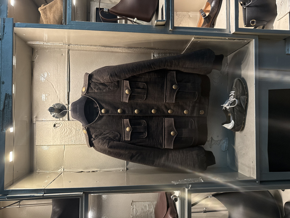 The scene depicts a meticulously arranged retail display within a high-end fashion boutique located in Paris, France, specifically a Dolce & Gabbana establishment as indicated by the brand on the garment hanger.
The central feature is a dark brown, long-sleeved jacket, suspended from a black plastic hanger. The jacket exhibits a military-inspired design with a stand collar, a row of eight gold-toned buttons down the front, and two pairs of patch pockets with gold-toned buttoned flaps on the chest and waist. Shoulder epaulets with single gold buttons and quilted shoulder panels further accent the garment. The sleeves have elasticated or gathered cuffs. Below the jacket, resting on a light gray shelf, is a single dark-colored sneaker, possibly black or dark gray, with white laces and a white heel tab.
The display unit is a multi-compartment structure, appearing to be constructed from distressed, off-white panels with visible seams and rivet-like details, suggesting an industrial aesthetic. Each compartment is individually lit by small, recessed square spotlights. Peripheral compartments partially visible include various leather goods: a black shoulder bag with a silver buckle, a dark brown wallet or clutch, another dark brown crescent-shaped shoulder bag, a tan-colored bag, and a pair of dark Mary Jane-style flat shoes. No individuals are present in the image, and no actions or interactions are occurring; the scene is static.