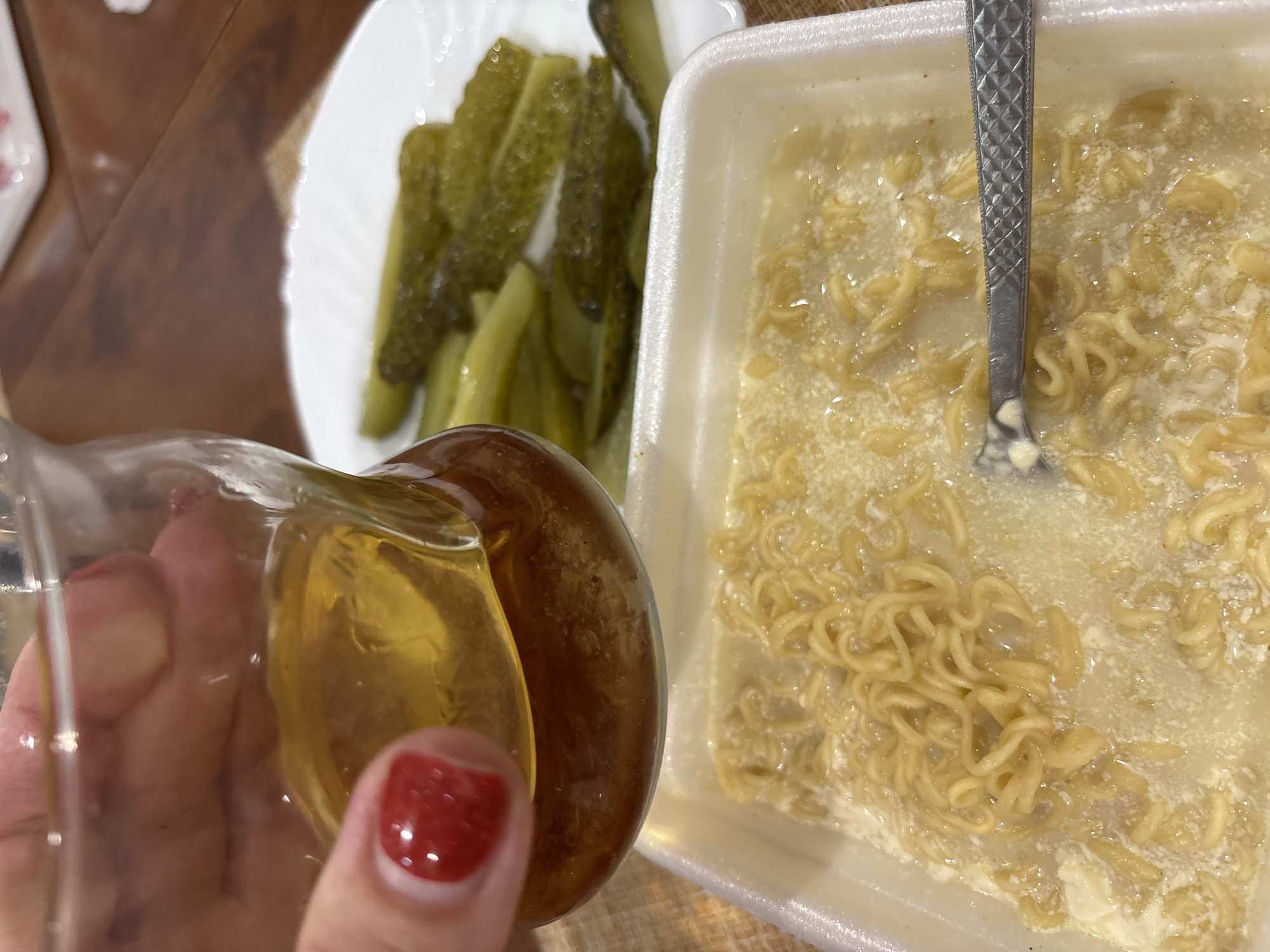 The image shows a person's hand with red nail polish holding a glass containing a golden-colored liquid. In the background, there are slices of pickles on a white plate and a white Styrofoam container filled with instant noodles in broth. A fork is visible in the container of noodles. The scene appears to be indoors, likely a casual meal setting. The lighting suggests it could be daytime. The presence of pickles and instant noodles suggests a simple meal. No text is visible in the image.