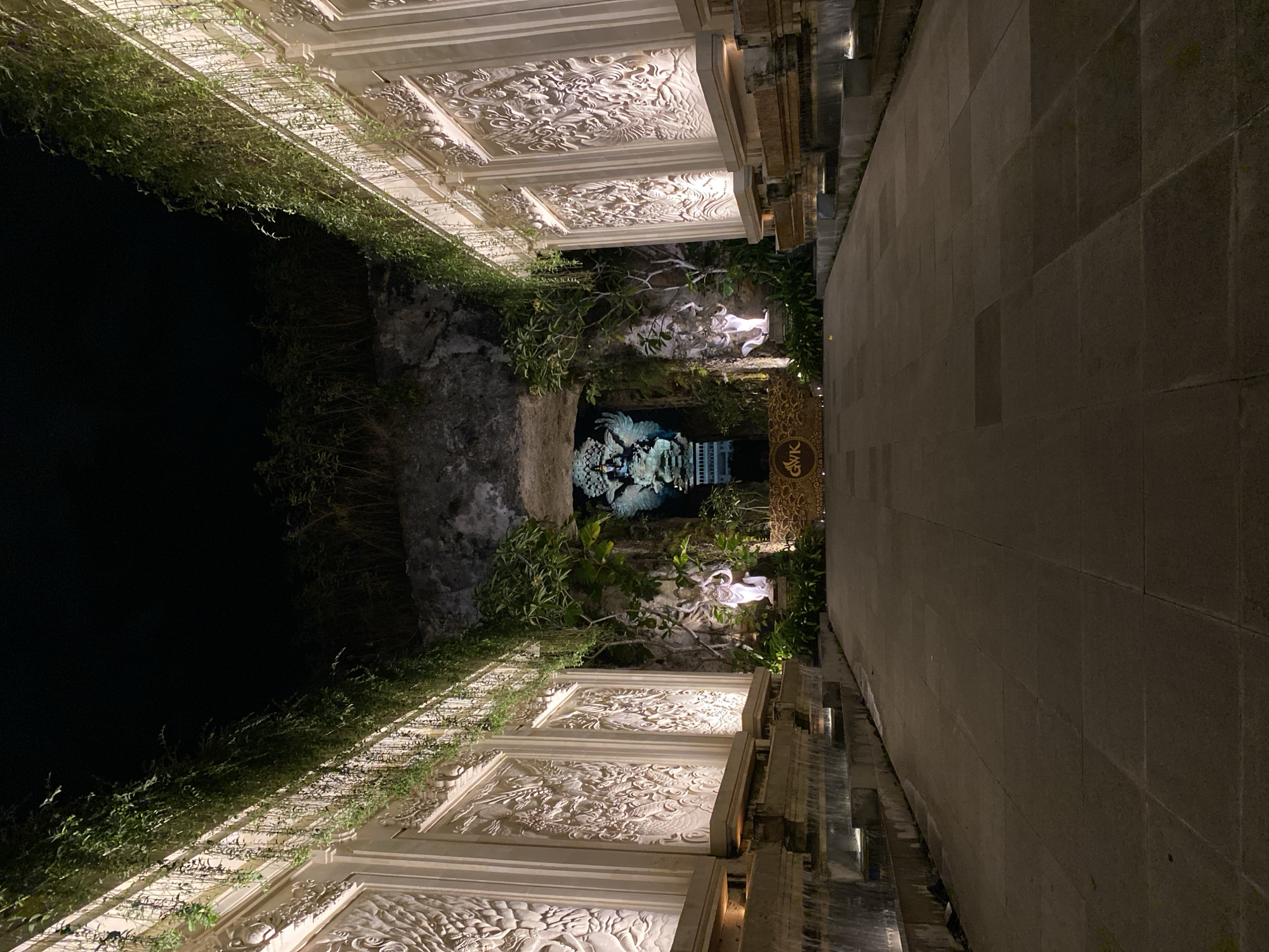 The image depicts a pathway at night, leading towards a large statue within a rocky alcove. The pathway is paved with large stone tiles and is flanked by ornate, carved stone walls on either side, illuminated by warm lights. Lush green vines cascade down from the top of these walls, creating a natural canopy overhead. At the entrance to the alcove, there are two smaller, illuminated statues. In the center of the alcove, a large, multi-armed statue, possibly of a deity, is visible, lit with blue and white light. A sign with the letters "GWK" is situated on the ground in front of the alcove. The overall scene is one of a well-lit, elaborately decorated outdoor space, suggesting a cultural or religious site.