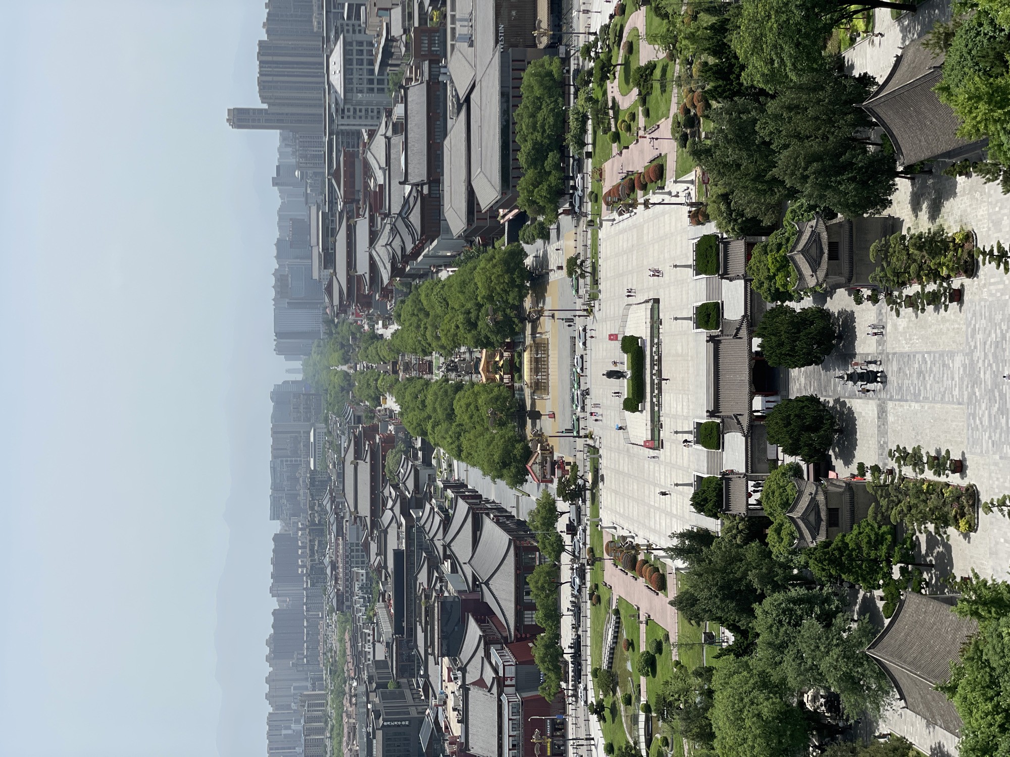 The image depicts an aerial view of a streetscape in Yanta District, China. It showcases traditional Chinese-style buildings with gray tile roofs aligned on either side of a wide pedestrian boulevard. Trees line the center of the boulevard, and smaller gardens are visible alongside it. In the foreground, parts of the street intersect at right angles. Pedestrians can be seen walking along the boulevard and other walkways. In the distance, modern high-rise buildings are visible. The sky is overcast.
