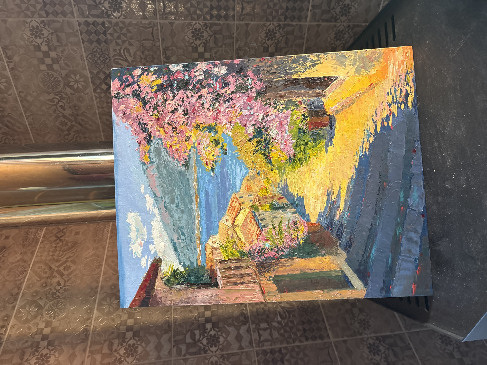 The painting depicts a sun-drenched, narrow street with buildings on either side leading down to a body of water. Lush pink and yellow foliage cascades from trees and potted plants. The architecture suggests a Mediterranean or Southern European coastal town. The foreground features dark, textured cobblestone steps dappled with sunlight. In the distance, a calm blue body of water is visible, bordered by distant hills. The sky is a bright blue with scattered white clouds. The painting is rendered in a thick, impasto style with visible brushstrokes, giving it a vibrant and textured appearance. The painting is placed in front of a wall with a tiled pattern and a metallic pipe running vertically. The overall impression is a picturesque and lively scene. There is no indication that this location is in Lyady, Russia.