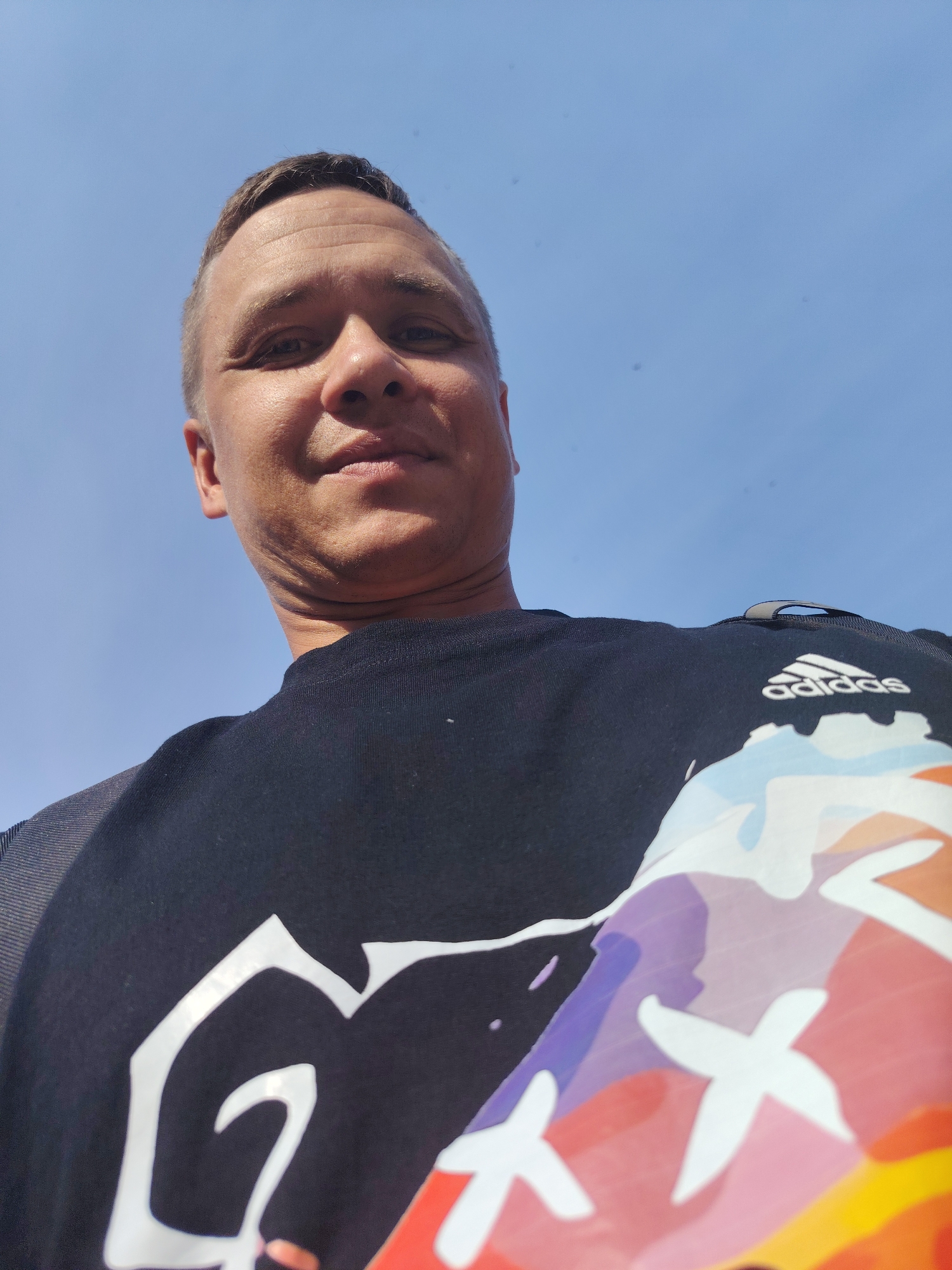 A low-angle, close-up, waist-up shot shows a Caucasian man with short, light brown hair and fair skin looking up at the camera. He is wearing a dark t-shirt with a colorful graphic design on the front, including white shapes and the Adidas logo. The man has a slight smile and wrinkles on his forehead and under his chin. The background is a clear, bright blue sky. The scene is outdoors, under daylight. There are no specific landmarks or environmental features visible that would indicate the location within the city of Chaikovsky, Russia.