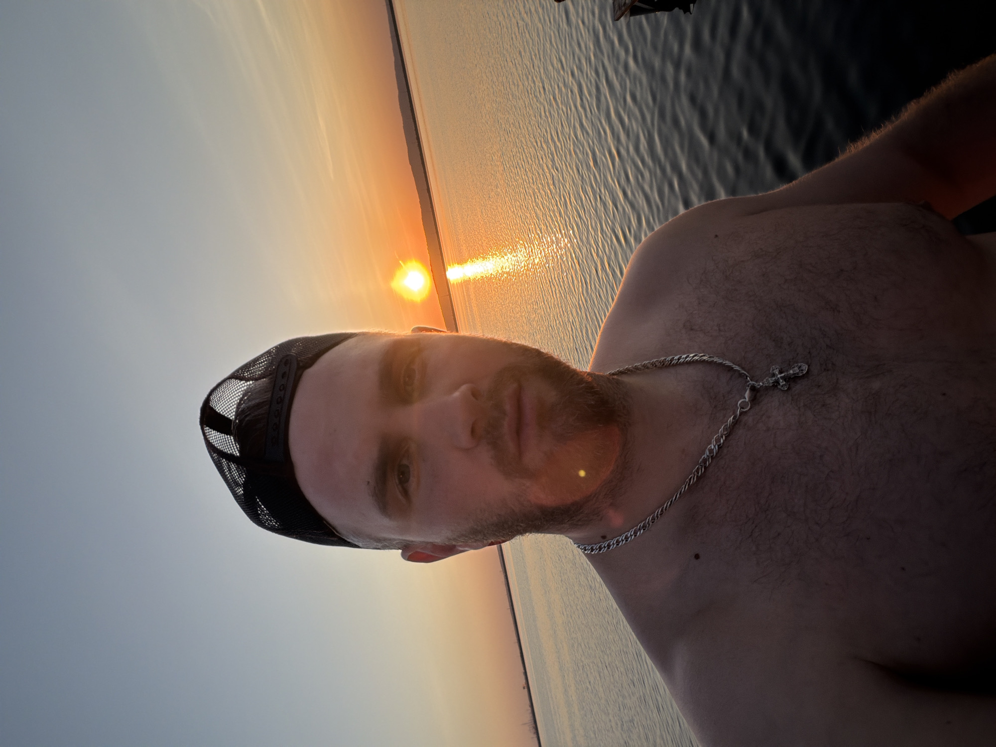 A medium shot depicts a light-skinned, bearded man from the chest up, wearing a black cap and silver necklace with a cross pendant. He stands on what appears to be a wooden structure over water, possibly a dock or pier. In the background, the sun sets over a body of water, creating a golden reflection. The location is likely in or near the city of Chaikovsky, Russia, given the prompt's instruction.