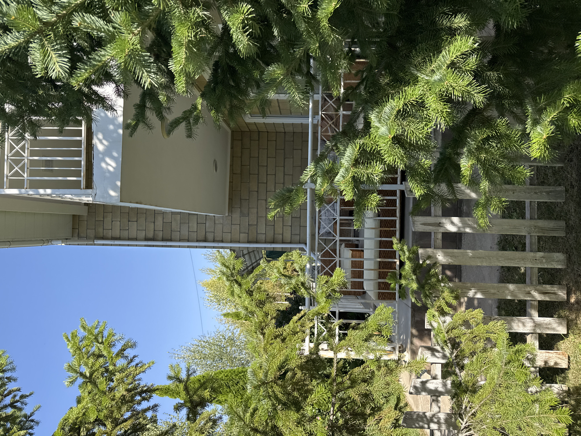 The image shows a residential building, likely a house, on a sunny day in Sary-Oy, Kyrgyzstan. In the foreground, lush green pine branches partially obscure the view. A white metal railing with decorative geometric patterns runs along a balcony on the second floor of the building. The building itself is constructed with light-colored bricks. Below the balcony, there are two wicker chairs with white cushions, suggesting an outdoor seating area. A low, weathered wooden fence stands in front of the building, separating it from the surrounding grassy area. The sky is a clear, bright blue. The overall scene depicts a peaceful residential setting with natural elements.