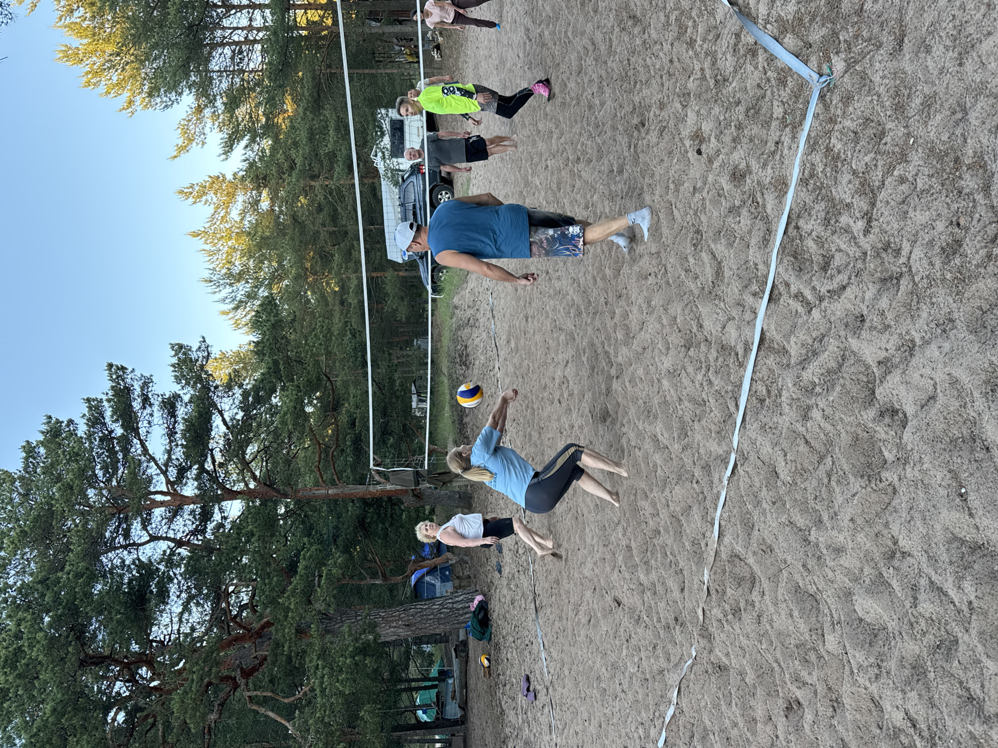 A group of people are playing beach volleyball on a sandy court surrounded by pine trees. The sky is clear and blue. In the foreground, a woman in a blue t-shirt and dark leggings is in the process of hitting a volleyball. To her left, another woman in a white tank top and black shorts is watching. Further back, several other people are visible, some standing and some appearing to be involved in the game. There is a volleyball net set up across the court. In the background, there are tents and a vehicle, suggesting a recreational area or campsite. The overall scene depicts a casual outdoor activity in a natural setting.
