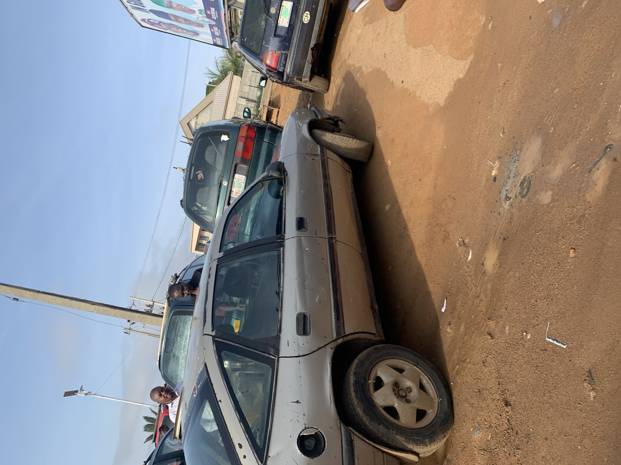 The image captures a bustling scene on an unpaved, dirt road in Rafin Par, Nigeria, under a bright, clear blue sky, indicating daytime. Several cars are present, suggesting traffic or parking along the roadside.
In the foreground, a silver or light grey four-door car is prominent, with its front right wheel severely angled outwards, indicating significant damage or a mechanical failure. Its body shows signs of wear and tear, including scuffs and a missing fuel cap cover. Another vehicle, a dark green car with a license plate "RNG-680XC" (a Nigerian registration), is parked behind it. Further back, a dark blue car is visible, its rear also showing some wear, and a partial license plate with "CH" visible.
Several men are visible in and around the vehicles. One man in a white shirt is seen leaning into the driver's side of the silver car, while another man looks out from the passenger window of the green car. Another individual is partially visible near the dark blue car. Their expressions are neutral, appearing engaged in the immediate surroundings.
The road surface is primarily dry, light brown dirt with patches of darker, possibly wet or muddy areas. Debris and small pieces of litter are scattered on the ground. To the left, a concrete utility pole with power lines runs vertically. In the background, low-rise buildings with light-colored walls and dark roofs are visible, along with some sparse vegetation like palm trees.
Above the vehicles, a large billboard is visible, partially obscured, displaying multiple faces and some indistinct text, suggestive of a political campaign or commercial advertisement. The only clearly legible text on a license plate is "RNG-680XC" on the green car. The overall impression is one of a busy local street with older vehicles, characteristic of a town or rural area in Nigeria.