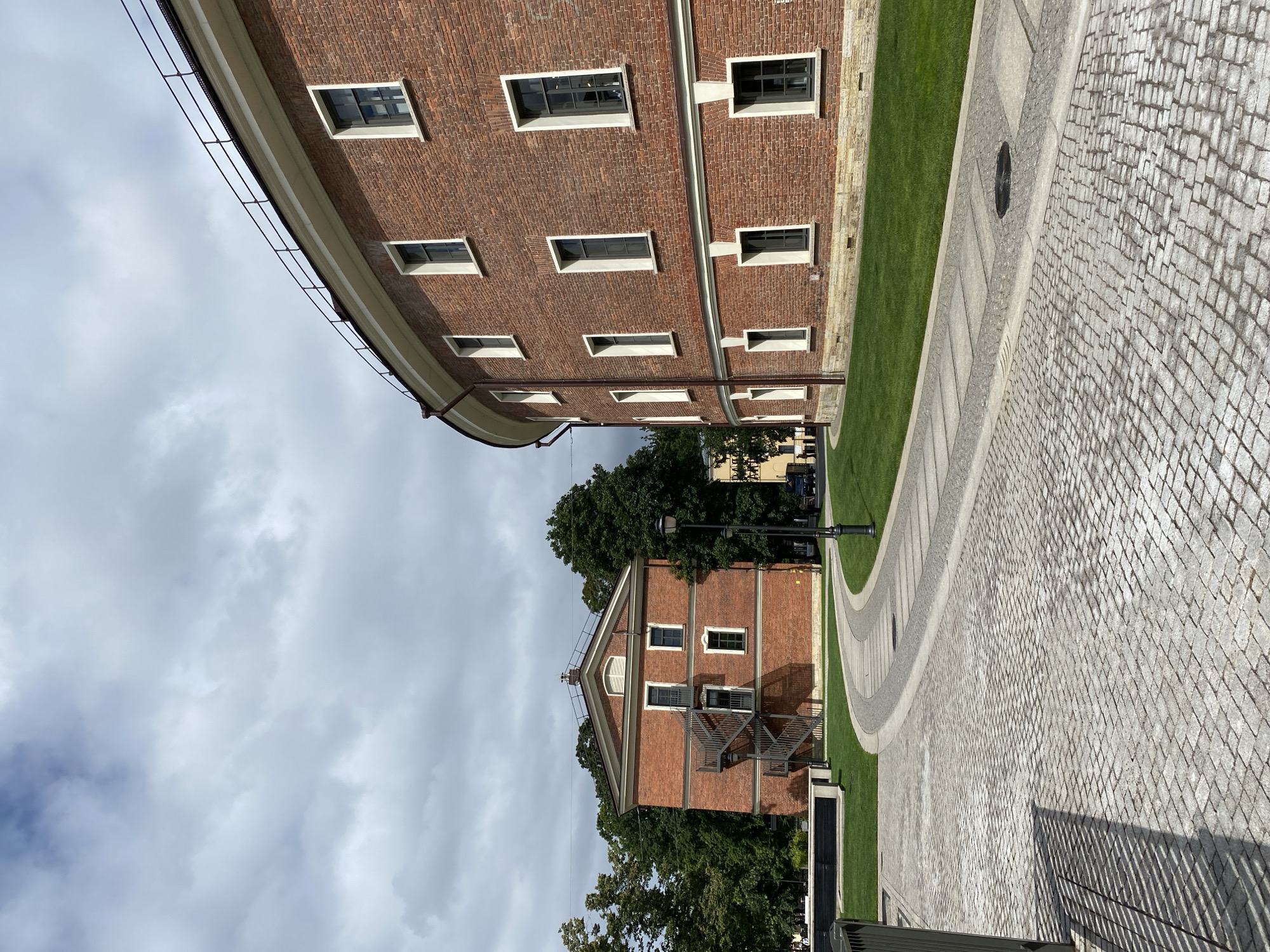 The image depicts an outdoor scene in Saint Petersburg, Russia, featuring two red brick buildings. The building on the right is curved, with multiple windows and a white cornice. The building on the left is rectangular with a pitched roof, an external metal staircase, and several windows. A cobblestone pathway curves through a manicured green lawn, leading towards the buildings. Trees surround the area, and the sky is partly cloudy. The scene is serene and architectural, with no people or vehicles prominently visible.