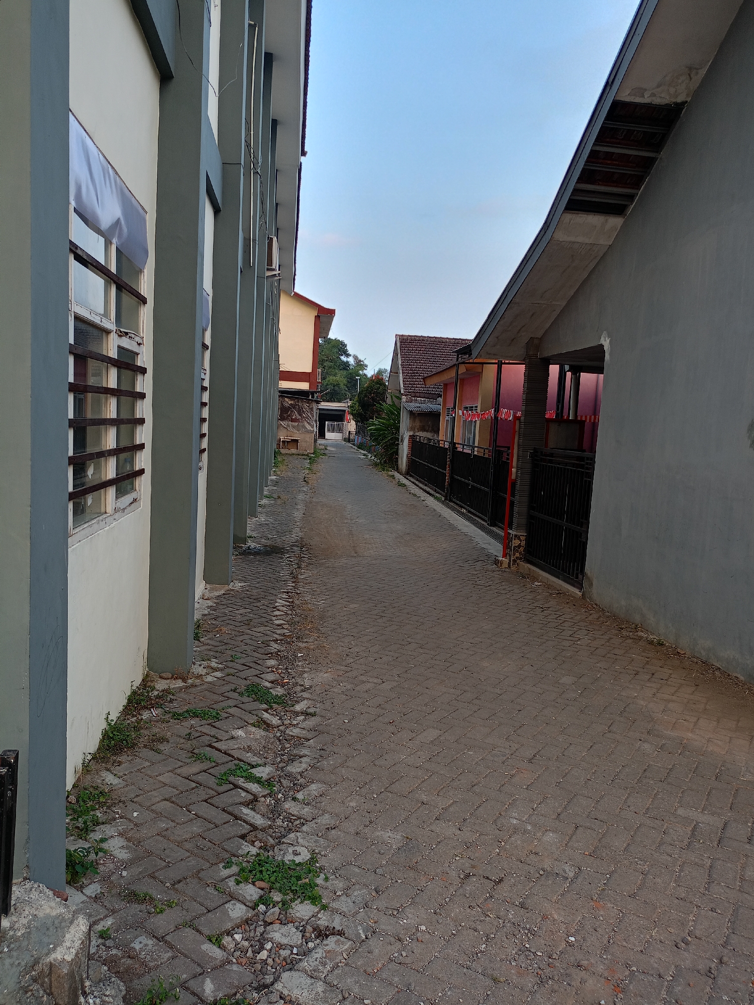 The image depicts a narrow, paved alleyway in the city of Batu, Indonesia, with buildings on either side. On the left, a light-colored building with grey vertical pillars and barred windows lines the alley. White fabric is visible behind some of the windows, partially obscuring the view. On the right, a grey building with a sloping roof dominates the foreground, with an opening suggesting a covered walkway or entrance. Further down the alley, residential buildings with tiled roofs and dark metal fences are visible. Some buildings are painted in warm tones like pink and yellow, with red and white decorative flags strung across the fence line, possibly indicating a local celebration or event. The alleyway is paved with interlocking grey bricks, some of which appear worn or displaced, with patches of grass and small stones growing in the cracks. The sky is clear and blue, suggesting daylight. The scene is devoid of people.