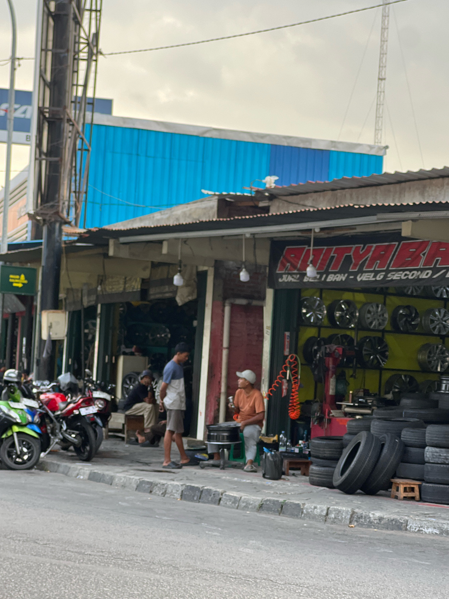 This image depicts a street-level view of a tire and wheel shop in the city of Yogyakarta, Indonesia. The shop appears to be an open-air establishment with a corrugated metal roof, situated along a paved road. In front of the shop, several motorcycles are parked, including a green, red, and white one. A stack of tires is prominently displayed on the sidewalk in front of the shop, and shelves inside are filled with various car wheels. Two men are visible outside the shop, one in a light-colored shirt and dark shorts, the other in an orange shirt and light-colored pants, seemingly engaged in conversation or observing the surroundings. Another individual is seated inside the shop. The background reveals a building with a blue corrugated metal facade and a tall communication tower in the distance. The overall scene suggests a busy commercial area with a focus on automotive services.