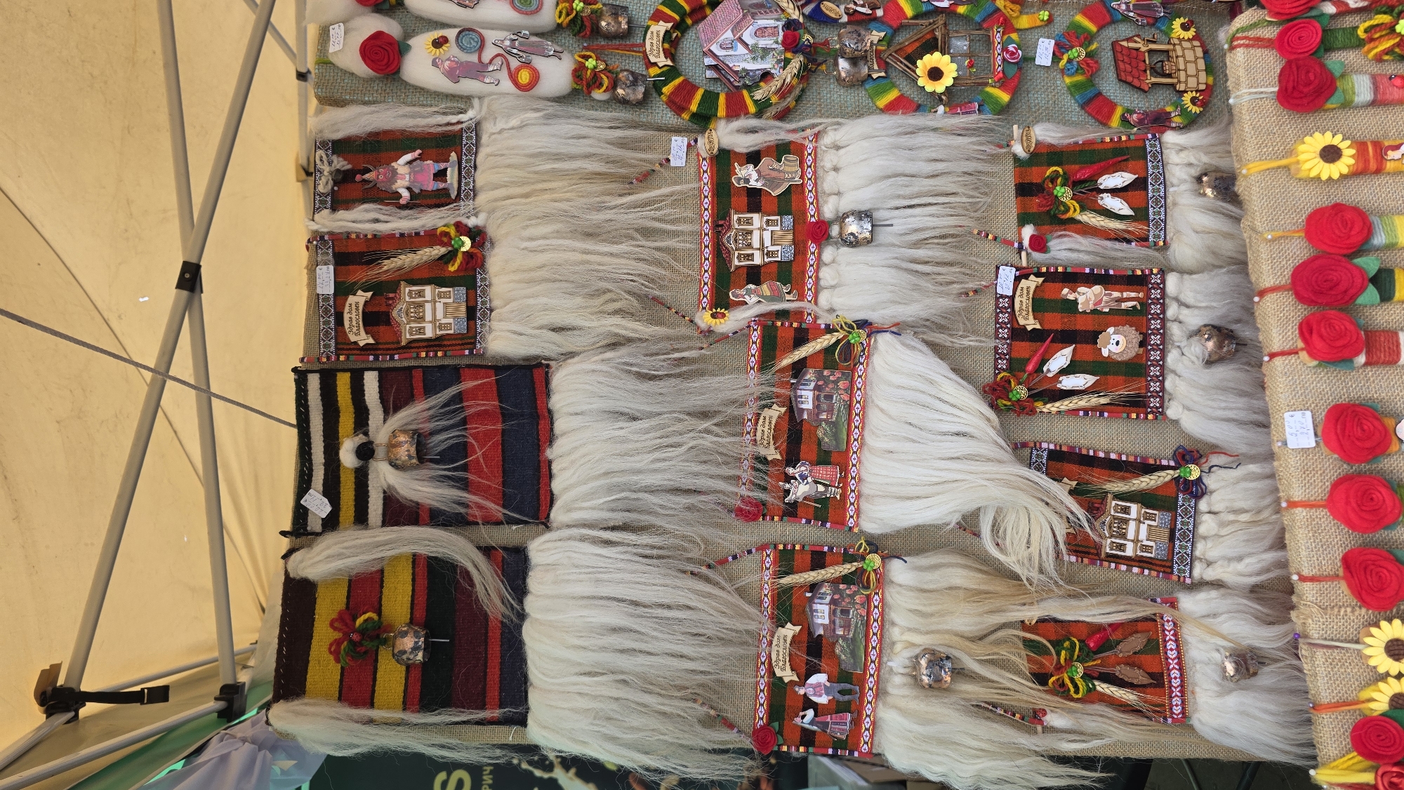 This is a top-down shot of a market stall in what may be Blagoevgrad, Bulgaria. On display are various handcrafted souvenirs laid out on a burlap-covered table. The items include: traditional Bulgarian "kukeri" masks with long, white faux fur, adorned with woven fabric squares and embellishments; felted wool ornaments shaped like slippers; and wreaths decorated with yarn, wooden cutouts of houses, and sunflowers. The stall is situated under a beige canopy, with parts of the metal frame visible on the left.