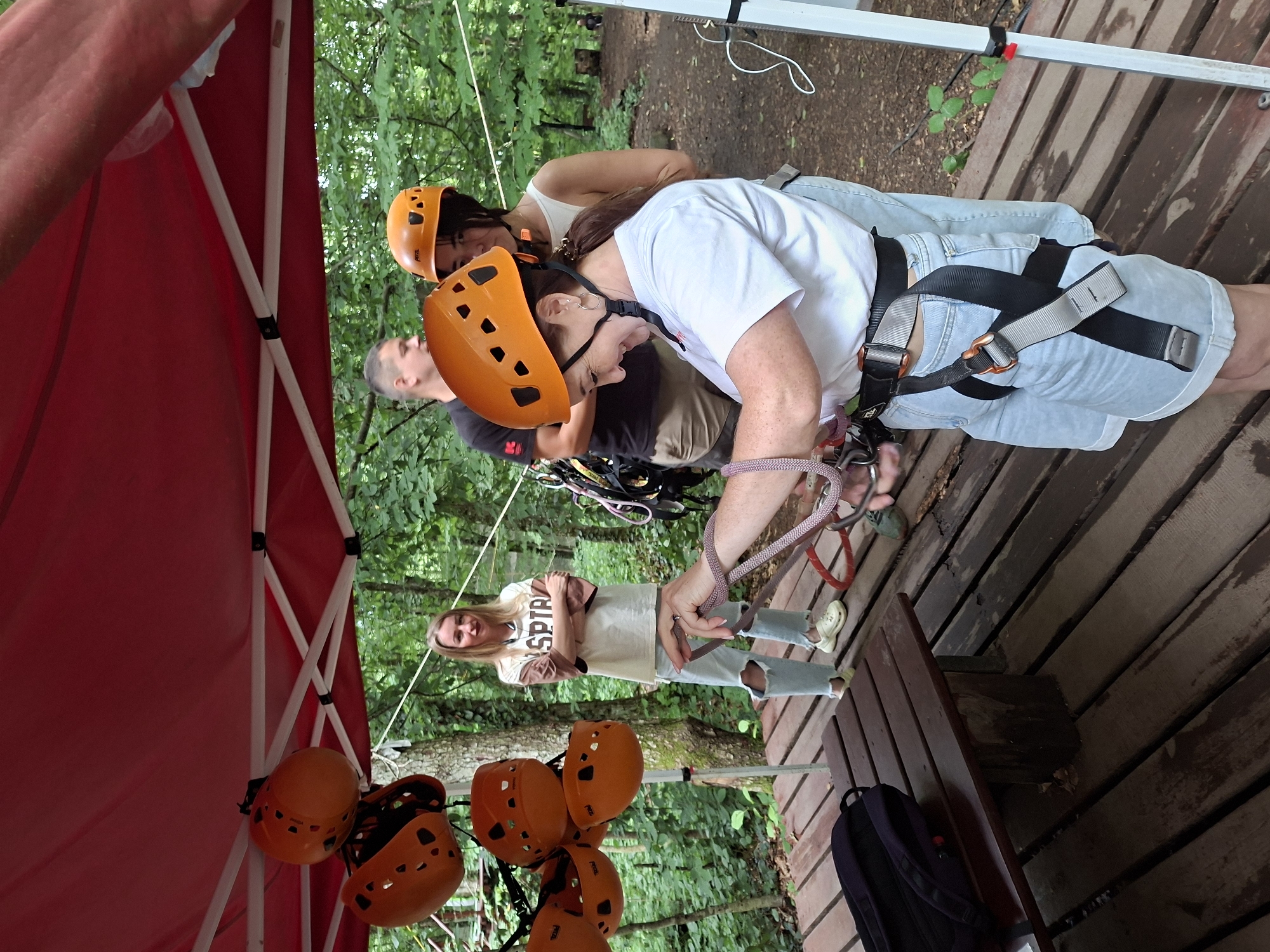 In the city of Unknown, Abkhazia, a group of people are preparing for an adventure activity under a red canopy. The scene is set outdoors, amidst lush green trees, suggesting a forest or park environment. A wooden platform serves as the ground for this area.
In the foreground, a woman wearing a white t-shirt, light blue denim shorts, and a helmet is actively engaged in handling climbing ropes and harnesses. She has a focused expression as she adjusts the equipment. Another woman, also wearing a helmet, stands behind her, observing. To the left, a woman in a light-colored t-shirt and ripped jeans stands with her arms crossed, looking towards the camera. In the background, a man in a dark t-shirt is also visible, appearing to be part of the group. Several orange helmets are suspended from the canopy, indicating the availability of safety gear. A dark backpack rests on a wooden bench on the left side of the image. The overall atmosphere is one of anticipation and preparation for an outdoor adventure.