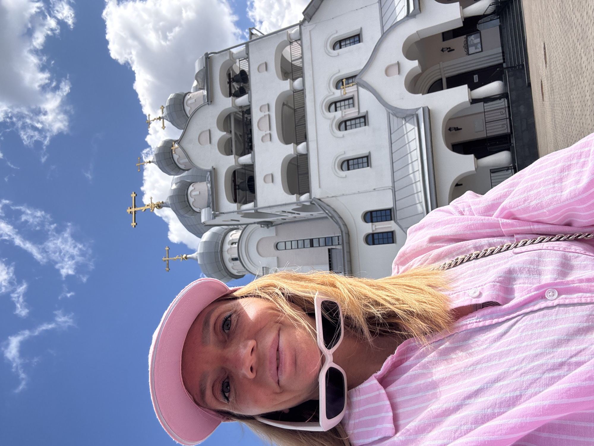 In this close-up, self-taken photograph, a woman stands in front of a white, ornate building featuring multiple domes and crosses, characteristic of Orthodox Christian architecture. She wears a pink baseball cap and a pink and white striped collared shirt, with a pair of white-rimmed sunglasses resting around her neck. The sky above is blue with scattered white clouds. The building, which appears to be a church or monastery, is a significant architectural element in the background, with visible windows, arched entrances, and several bells hanging in an upper belfry. The ground in the foreground is paved with light-colored square stones. This scene is set in Saint Petersburg, Russia, at the Church of the Resurrection of Lazarus within the Alexander Nevsky Lavra.