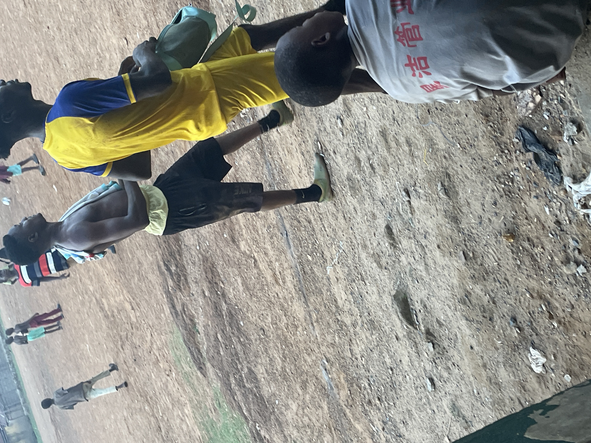 This media file depicts an outdoor scene in Akwanga, Nigeria, under bright daylight. The primary subjects are several young, dark-skinned individuals gathered on a dusty, reddish-brown unpaved ground that appears to be an open field or training area.
In the foreground, three young men are prominent. One on the far right wears a grey t-shirt with red Chinese characters, which clearly include "福音" (Fúyīn, meaning Gospel or Good News). He is seen from behind, facing away from the camera. Next to him, another individual wears a yellow and blue athletic t-shirt, yellow shorts, and has a light green bag slung over his shoulder. He is standing, facing towards the left side of the frame. On the left side of the foreground, a third young man is shirtless, wearing black shorts with a light-colored cloth draped over his shoulder, also facing left. His shorts feature a "NIKE" logo.
In the mid-ground and background, several other individuals, some wearing t-shirts and shorts, are visible standing informally on the unkempt ground. The attire of the individuals suggests a casual or sports-related activity, possibly a training session or a general gathering. The ground is uneven, with scattered debris and small patches of green vegetation. The scene conveys a relaxed and informal atmosphere.