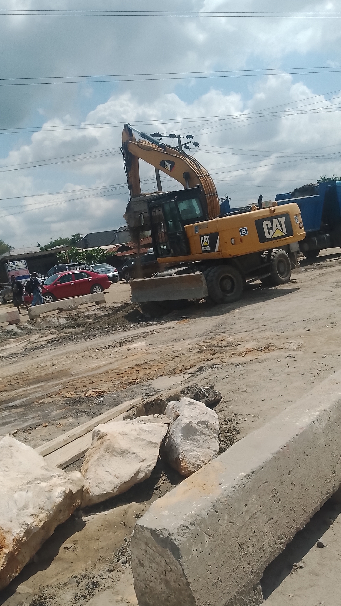 The image shows a construction site in Effurun, Nigeria. A yellow CAT excavator is the focal point, with its arm raised. Nearby are several large, irregular white rocks and a long, rectangular concrete block. In the background, there are people, vehicles including a red car and a blue truck, and buildings, suggesting an urban setting. Power lines are visible in the sky. The ground is uneven and appears to be dirt and gravel.