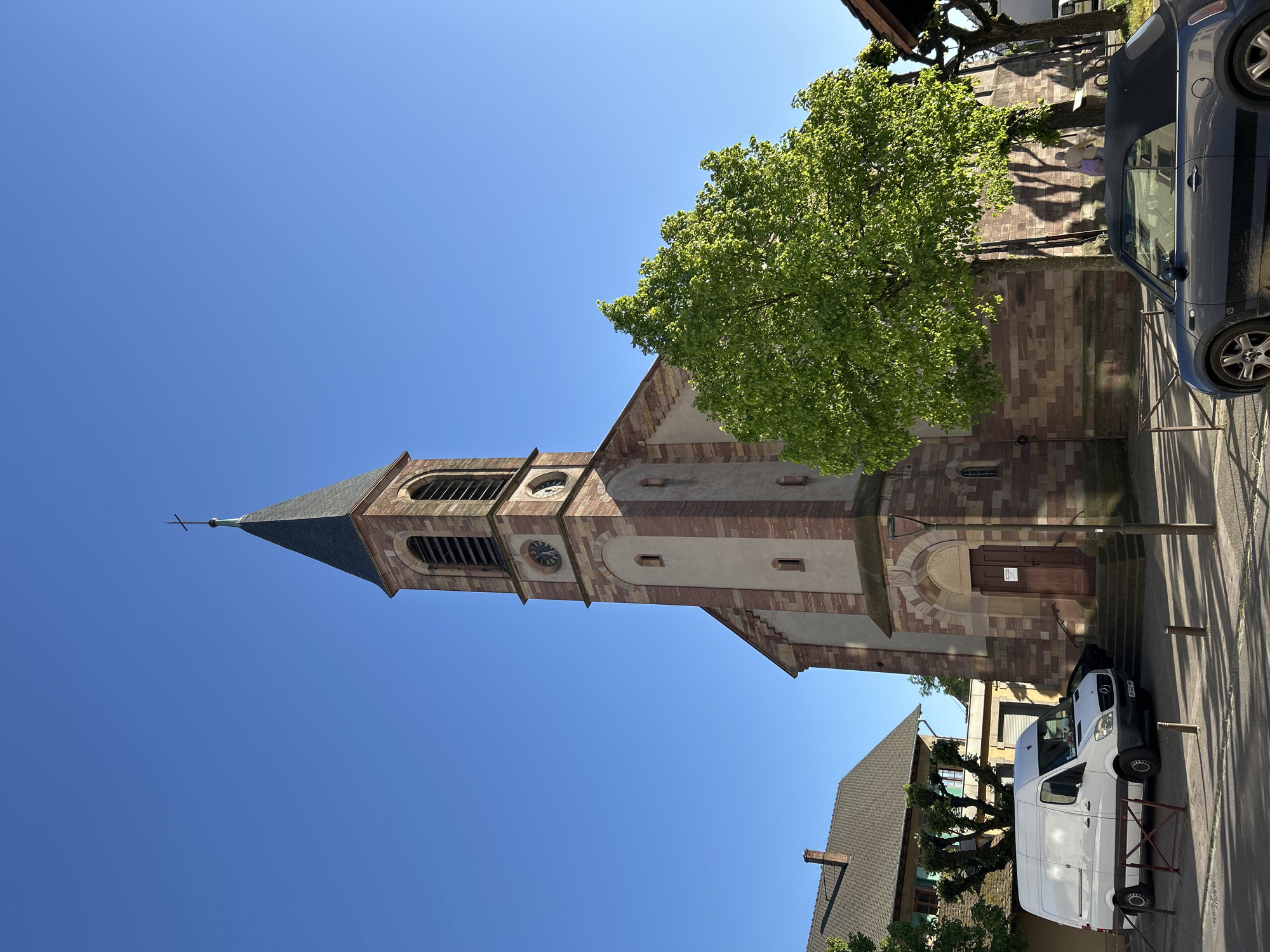 This media file captures a sunny, clear day in what appears to be a European town or village, identified as Strasbourg, France. The central subject is a stone church featuring a prominent bell tower with a pointed steeple, topped by a cross. The church walls are constructed from reddish-brown stone blocks interspersed with lighter-colored plaster sections. A clock face is visible on the tower, which also has arched openings for bells. The church's main entrance consists of wooden double doors, with steps leading up to it.
To the right of the church, a large deciduous tree with vibrant green foliage is visible, casting long shadows across the ground and nearby structures. Two vehicles are parked on the paved area: a dark blue convertible car on the right side of the frame and a white delivery van, displaying the license plate "BN-344-MM", nearer to the church's entrance. Several residential buildings with tiled roofs are partially visible in the foreground and background. The bright blue sky is cloudless, and the angled shadows suggest it is either morning or late afternoon. There are two unreadable informational signs on the church door. No people are visible, and no specific event is taking place; the image depicts a static street scene.