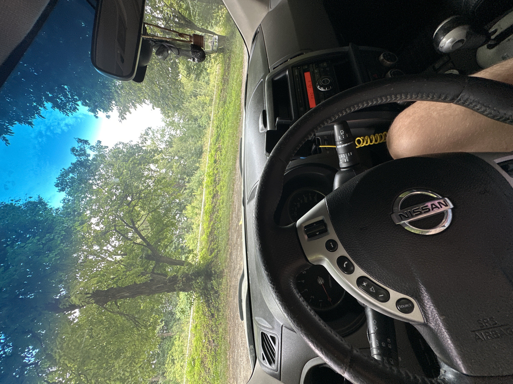 The image shows the interior of a Nissan car parked on a dirt road, with trees and vegetation visible through the windshield. The dashboard, steering wheel, and gear shift are prominent. A leg is visible through the steering wheel, suggesting a person is in the driver's seat. An air freshener hangs from the rearview mirror. The sky is blue with some clouds. The location is indicated as Shatava, Ukraine.