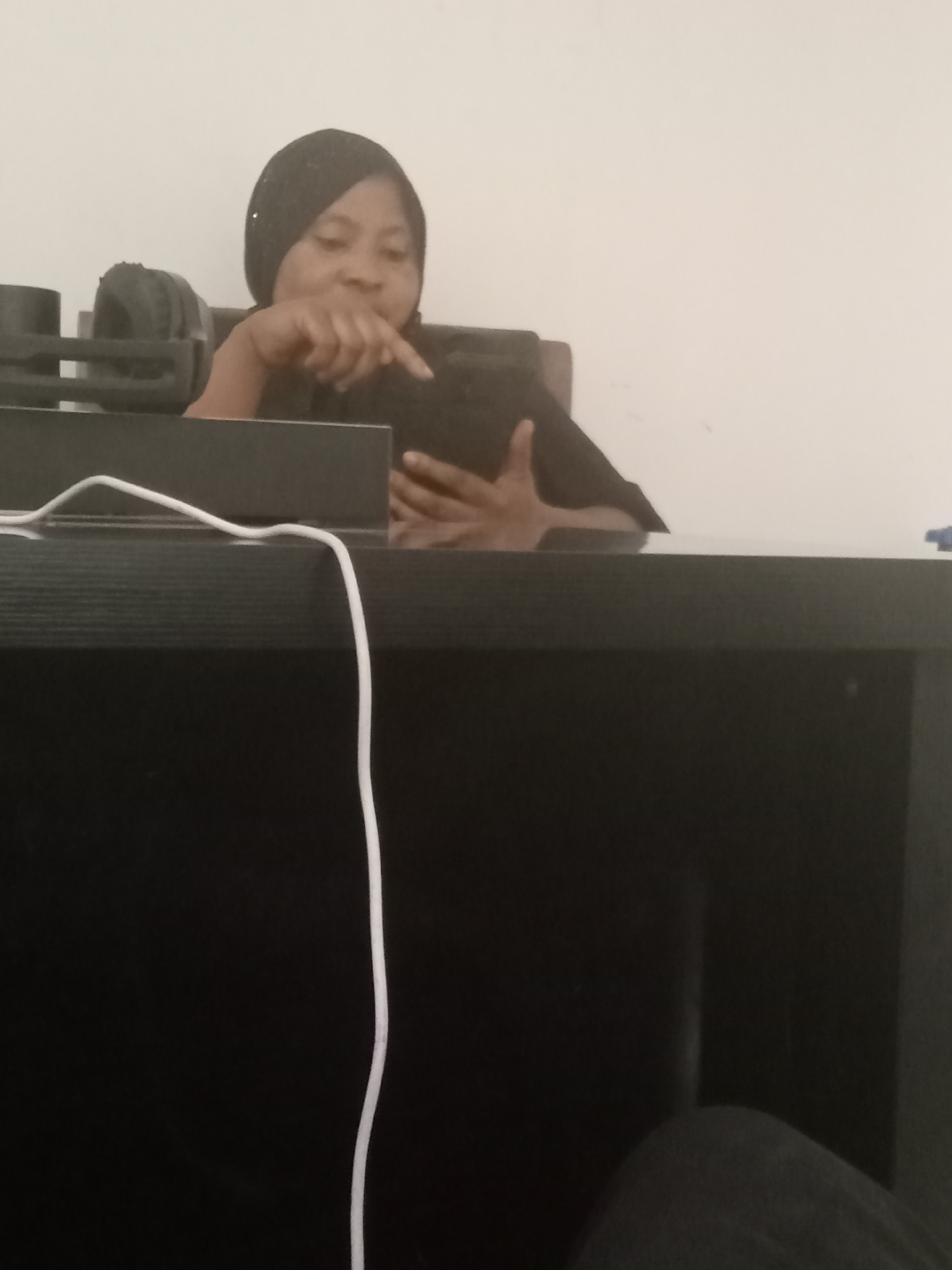 A woman wearing a dark headscarf is seated behind a dark-colored desk, looking at a device she is holding. Her right index finger is extended towards the device's screen. Headphones are seen in the background. A white cable is visible on the desk's surface. The background is a plain white wall. The photo's location is Gwarinpa, Nigeria.