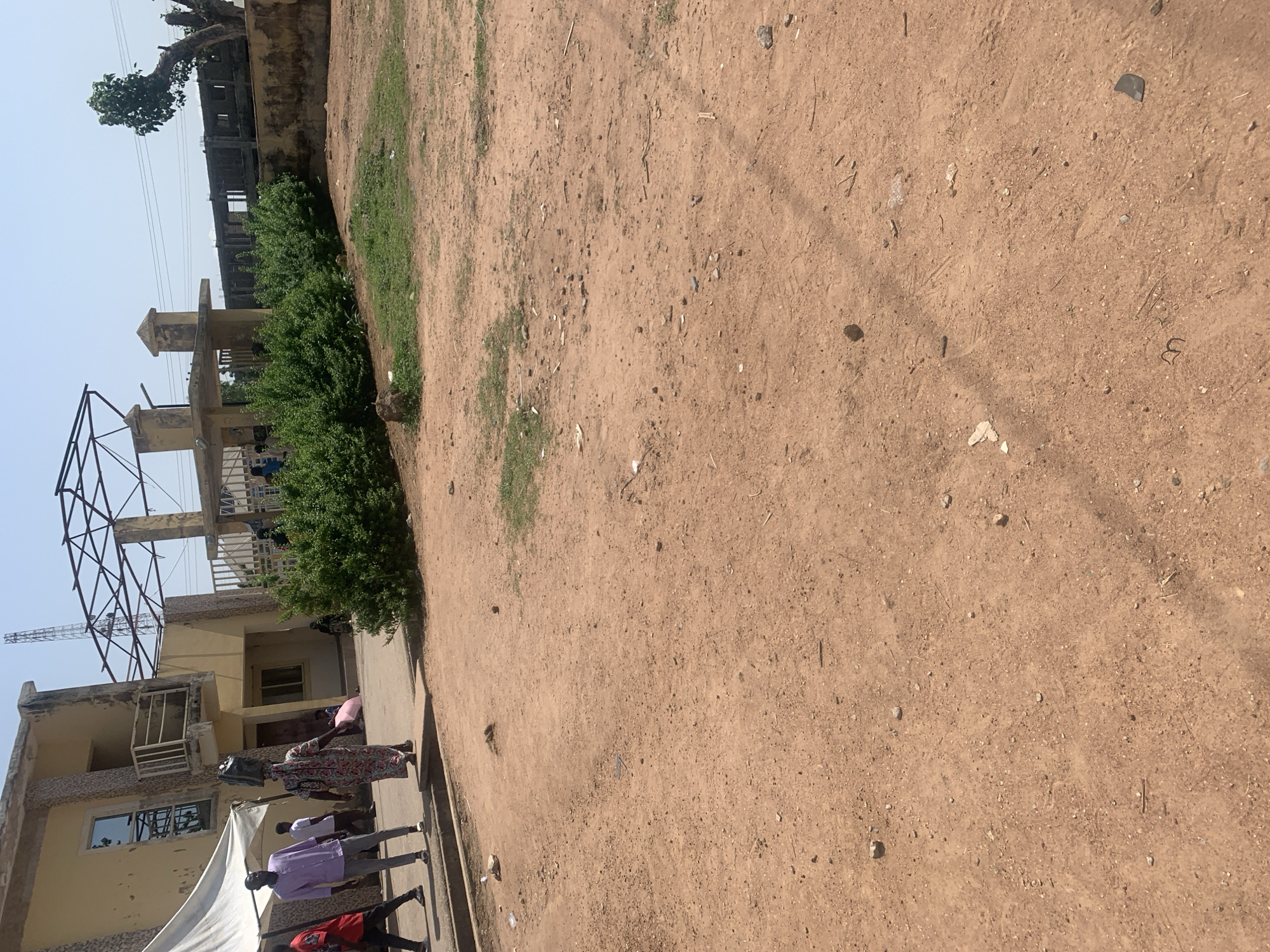 The image depicts an outdoor scene in Jalingo, Nigeria. The foreground is dominated by a dirt and gravel ground, with some sparse patches of dry grass. In the background, a multi-story building is visible, appearing to be under construction or in a state of disrepair. The building has a light yellow facade with some stone detailing. A metal frame structure, possibly for signage or scaffolding, juts out from the side of the building.
A group of people are gathered near the building, walking along a paved area. One person wears a purple shirt, another a patterned dress, and someone else a red shirt. The overall atmosphere suggests a daytime scene under clear, bright sky, typical of a sunny day. The presence of construction elements and the general state of the buildings suggest a developing or perhaps economically challenged area. No text is clearly visible in the image. The scene appears to be a street or an area adjacent to a commercial or residential building. The people's postures do not convey any strong emotions, suggesting a casual movement through the area.