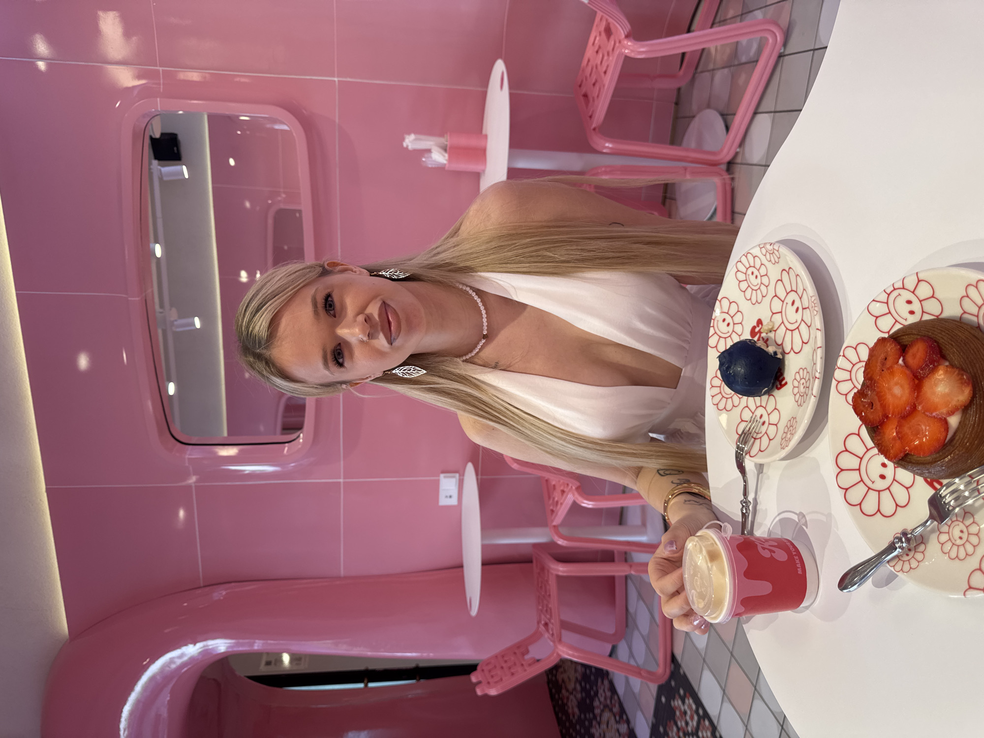 A young woman with blonde hair sits at a white table in a pink-themed establishment. She wears a white dress with a plunging neckline and pearl jewelry. In front of her are two plates with flower designs, one holding a blue dessert and the other a strawberry tart. A small red cup with a lid sits to her left. The room features pink walls, chairs, and architectural elements. The flooring is tiled with a grey and white checkerboard pattern. The establishment is located in Saint Petersburg, Russia.