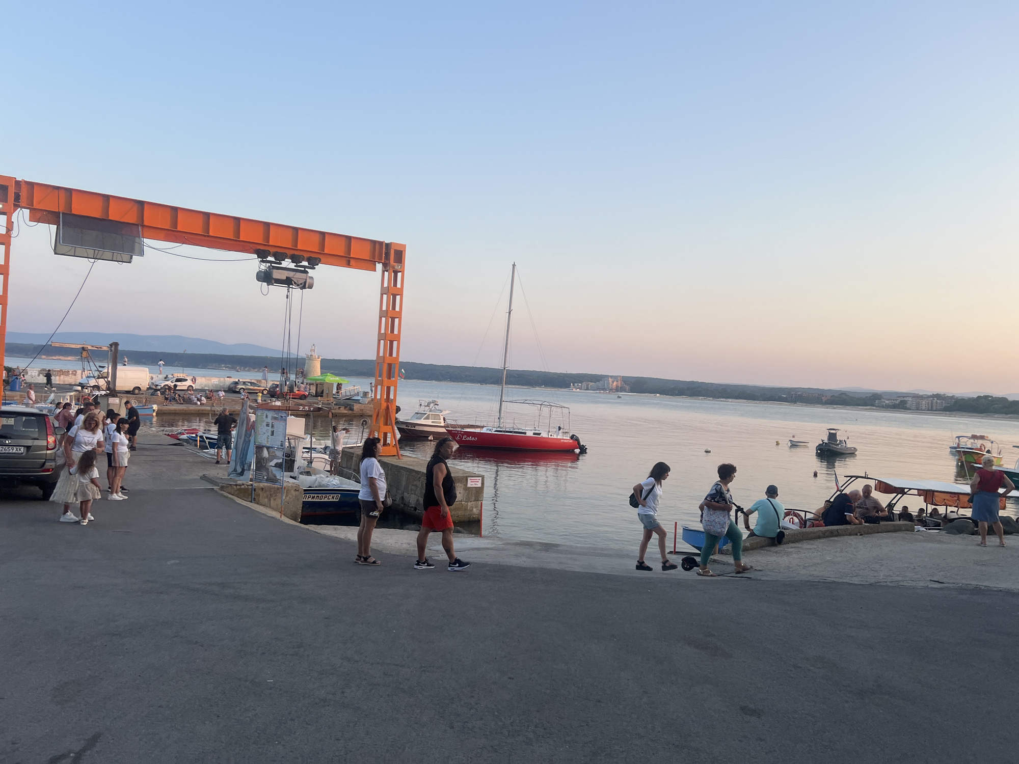 The image depicts a harbor scene in Primorsko, Bulgaria, during what appears to be sunset. A large, orange gantry crane dominates the left side of the frame, with a screen mounted on its horizontal beam. Several people are visible on the paved waterfront, some standing, others walking. A woman in a white top and denim shorts walks to the right, carrying a backpack. Beside her, a woman in a patterned top and green pants walks with a shopping cart. Further to the right, a group of people are gathered around a boat, some seated, others standing. Boats of various sizes are docked in the water, including a prominent red sailboat with "El Lotus" written on its side. The water is calm, reflecting the soft colors of the sky, and a distant shoreline with buildings and vegetation is visible. The overall atmosphere is relaxed, suggesting a leisurely evening at the port.