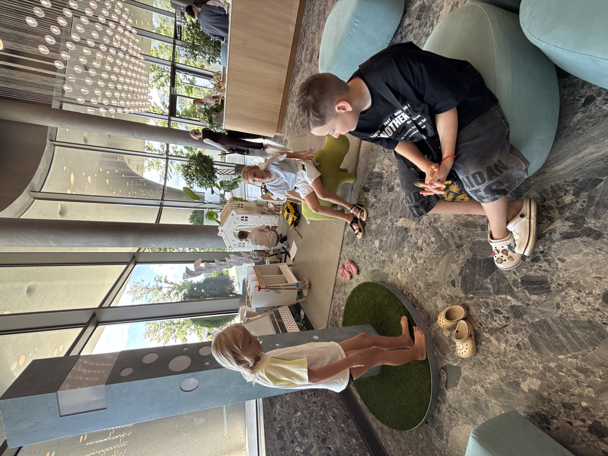 This image depicts an indoor play area, likely within a public establishment such as a hotel or restaurant, as suggested by the modern architecture and the presence of staff members in the background. The flooring is a polished grey marble.
In the foreground, a young boy with blonde hair, wearing a yellow t-shirt and black shorts, stands barefoot on a circular patch of artificial grass. Next to him, a pair of tan clogs and pink flip-flops are scattered on the floor. To the right, a young boy with short brown hair is seated on a light blue ottoman, wearing a black t-shirt and dark grey shorts. He has a small yellow bag across his chest.
In the mid-ground, a young girl with blonde hair is sitting on a green, hippo-shaped seat. She is wearing a light blue t-shirt and white shorts. Beside her, a yellow toy car is on the floor. Behind them, a white toy house and a wooden play kitchen are visible, suggesting a dedicated children's play zone. Further in the background, several adults, some appearing to be staff, are standing behind a wooden counter, interacting with the environment. Large windows offer a view of trees and the sky, indicating an outdoor setting beyond the interior. The overall atmosphere is casual and family-friendly. There is no indication that this location is in the city of Голубинское сельское поселение, Ukraine.