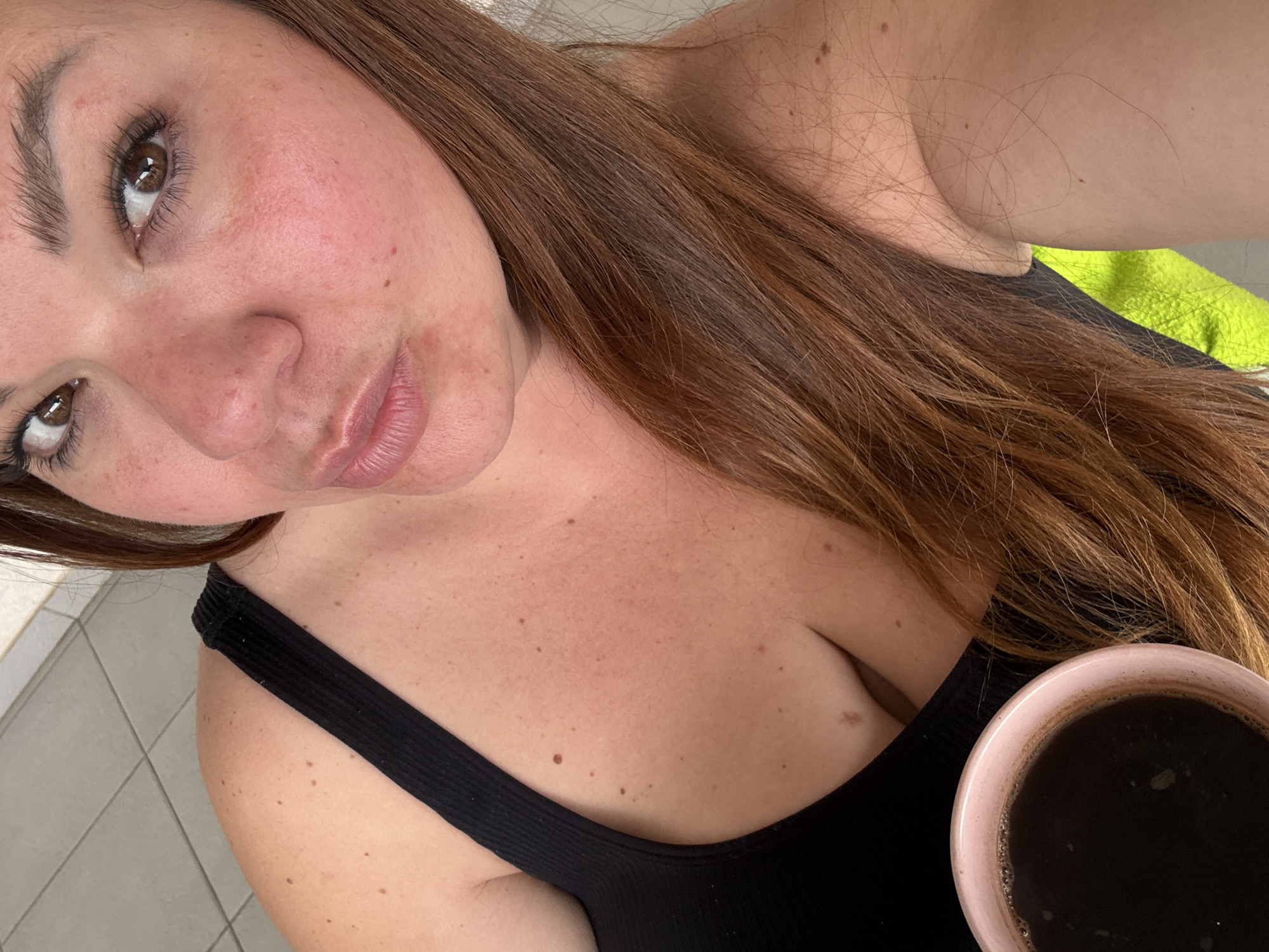 A woman with light skin, brown hair, and brown eyes is holding a mug filled with a dark liquid. She is wearing a black tank top. Her skin has various moles and minor skin blemishes. A section of wall with light-colored tile is visible. The location is the city of Bijela, Montenegro.