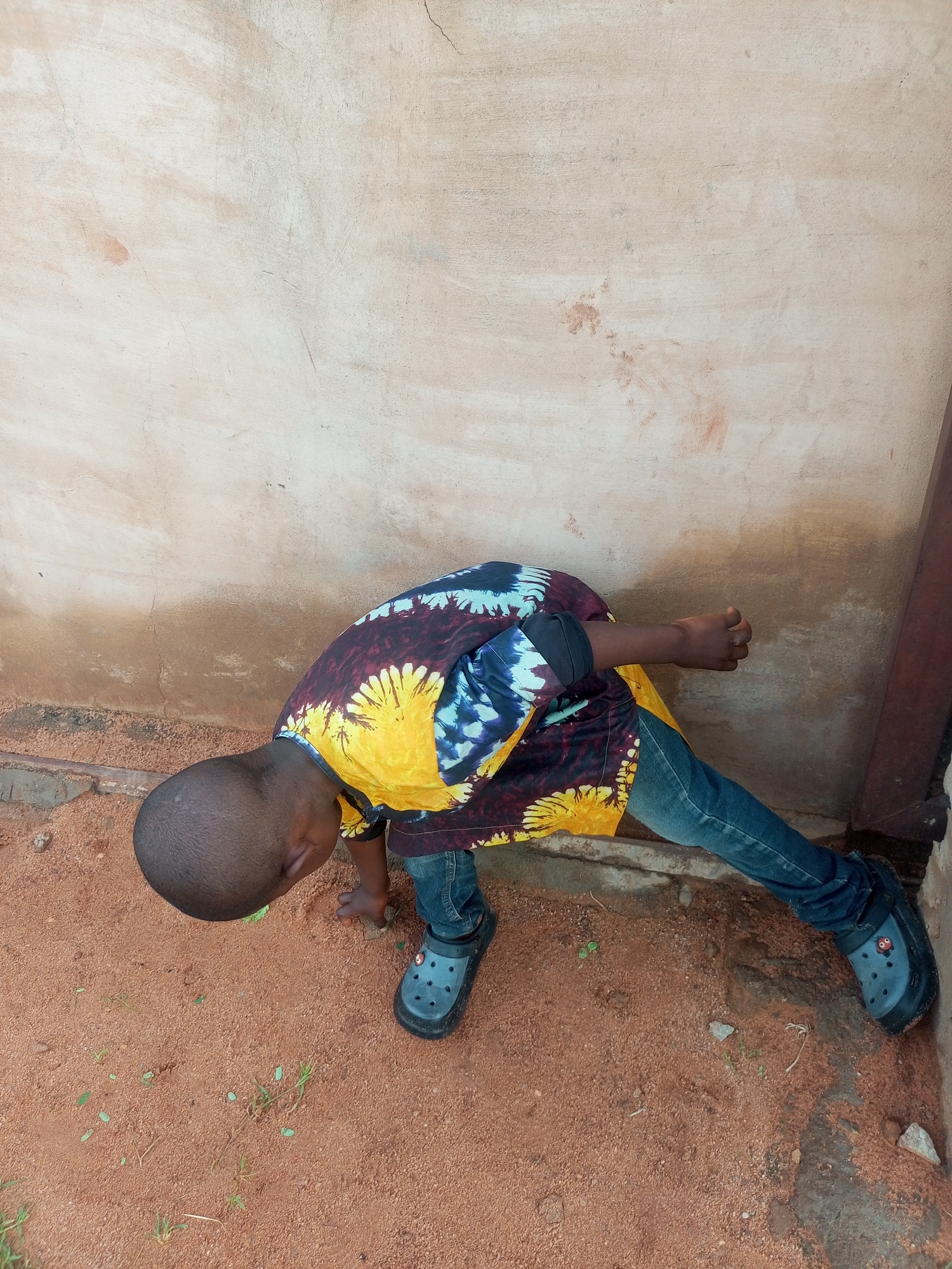 A young boy, appearing to be of African descent and with a shaved head, is bent over in a sandy, reddish-brown outdoor environment, possibly a courtyard or yard, near a textured, beige-colored wall. He is wearing a colorful, tie-dye patterned shirt with prominent yellow, blue, and black elements, blue denim jeans, and dark gray Crocs-style shoes. The boy is focused on something on the ground, with his left hand outstretched and his head down. The ground is covered in fine sand with scattered small pebbles and sparse green blades of grass. To the right of the boy, a vertical, dark metal structure, possibly a pipe or support beam, is visible against the wall. The wall itself shows signs of age and some discoloration, with a reddish-brown smudge near the bottom. The scene suggests a casual, everyday moment in an outdoor setting.