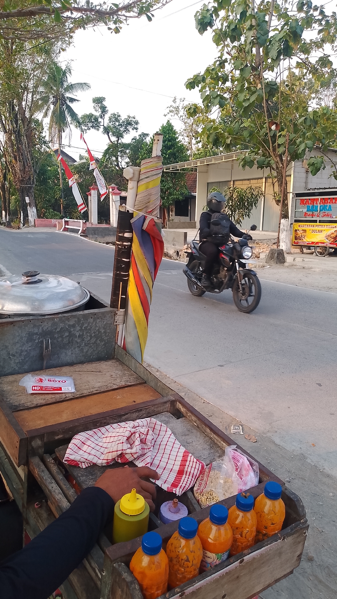 In the city of Karanglangu, Indonesia, a street scene unfolds with a food cart in the foreground and a street in the background. The food cart, made of weathered wood and metal, displays various items. A large, lidded pot sits on the left side of the cart. In the center, a red and white checkered cloth is folded, and a hand with dark blue sleeves reaches towards it. Beside the cloth, there are condiment bottles: one green with a yellow cap, a smaller purple-lidded container, and several orange bottles with blue caps. A bag of dry goods is also visible.
To the right of the cart, a person wearing a black outfit and a helmet rides a black motorcycle. The motorcycle is in motion, on the paved road. Behind the motorcycle, a white building with a sign that reads "MARTABAK BALIGKA" is visible. Indonesian flags, red and white, are displayed on poles in the middle ground. The environment is characterized by trees, a paved road, and a sidewalk, suggesting a typical Indonesian street scene. The lighting indicates it is late afternoon or early evening.