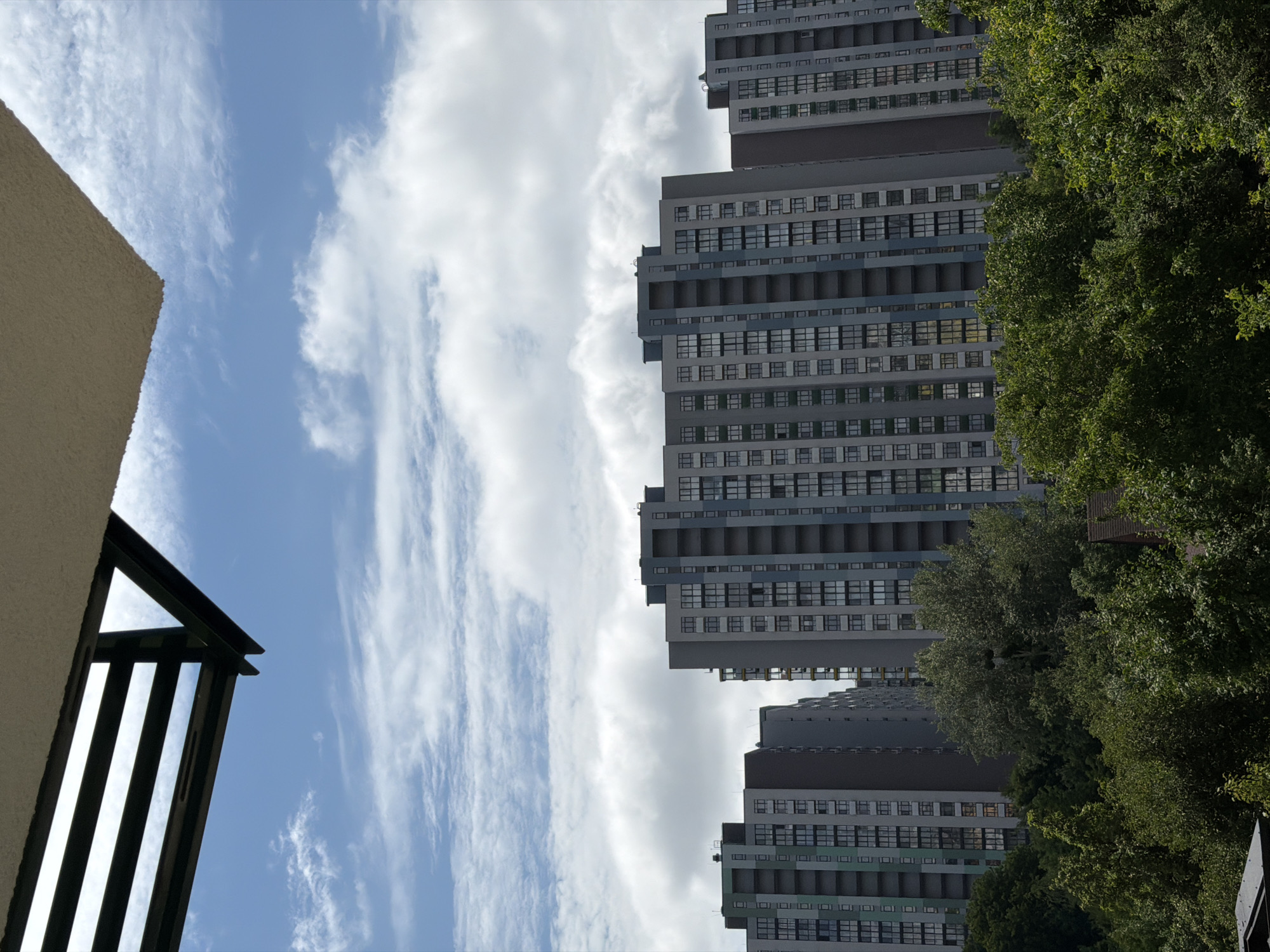 The image shows a view from an elevated position overlooking a residential area in Kyiv, Ukraine. In the foreground, a section of a building's overhang with a dark metal structure is visible against a blue sky with scattered white clouds. Below the overhang, several tall, modern apartment buildings dominate the mid-ground. These buildings are characterized by their grey facades, numerous windows, and balconies. Lush green trees and foliage fill the lower portion of the image, partially obscuring the lower levels of the apartment buildings and suggesting a well-treed urban environment. The scene depicts a typical urban landscape, with a mix of residential architecture and natural greenery.