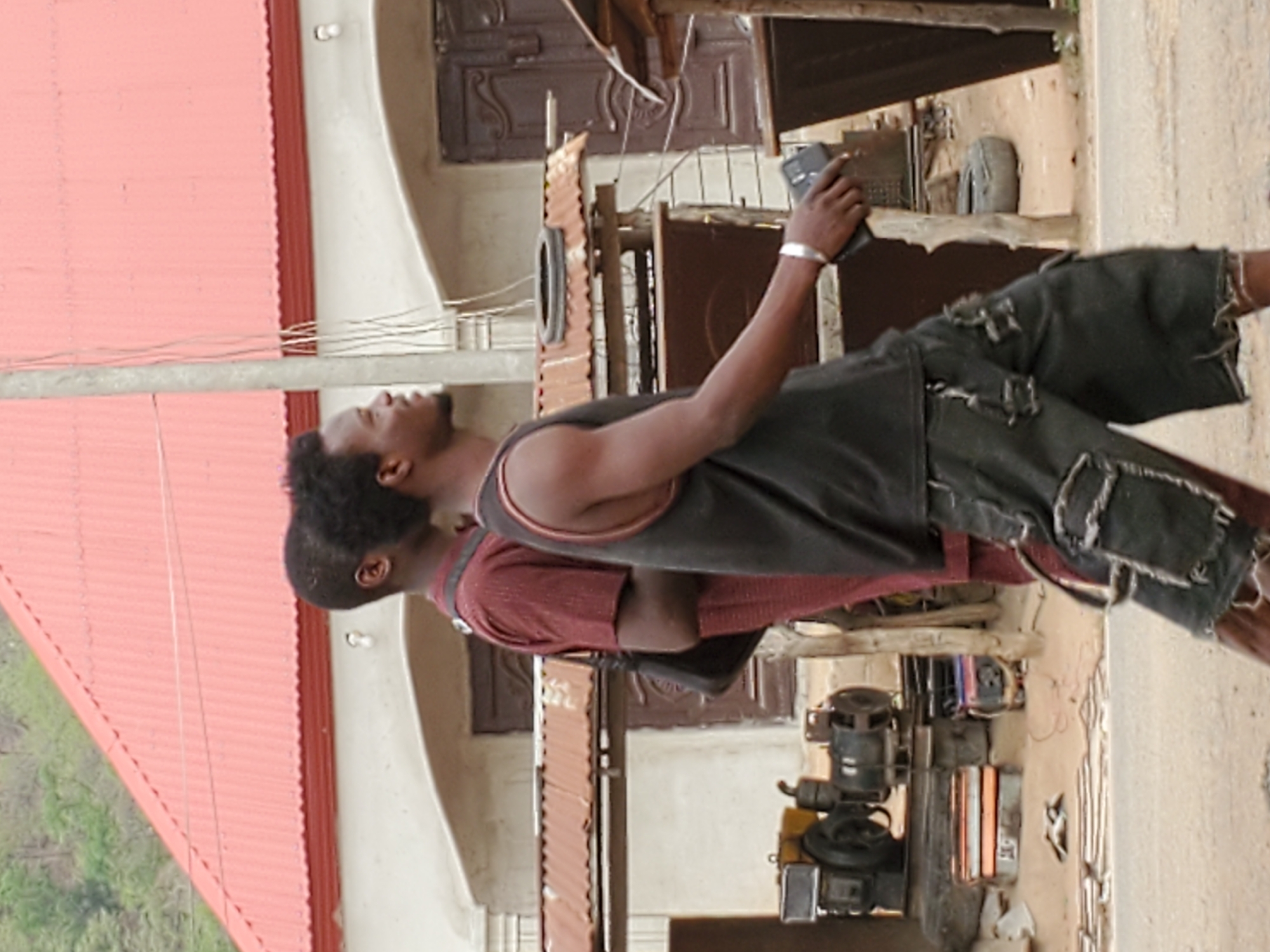 Two men are standing side-by-side, one holding a mobile phone. Both men are dark-skinned, and one is wearing a grey tank top over a maroon t-shirt and distressed denim shorts. The other man is wearing a maroon t-shirt. They are located outdoors near a corrugated metal roof, a concrete wall, and what appears to be a generator. The specific location is inferred to be in Mararaba, Nigeria.