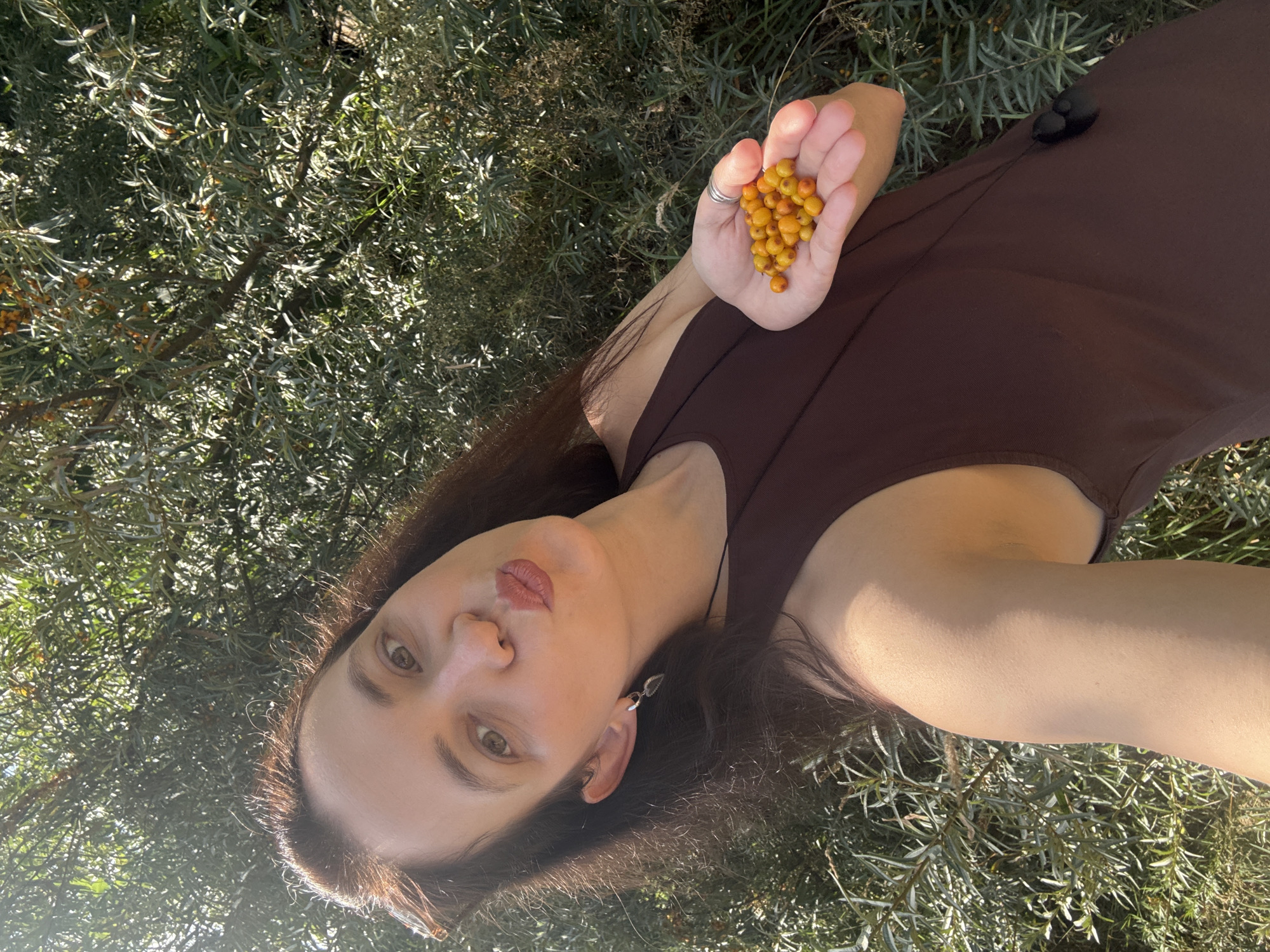 A woman with long, dark brown hair, wearing a dark brown sleeveless top and silver earrings, is holding a handful of small, round, orange berries in her left hand. She is looking towards the camera with her lips pursed. She is outdoors, surrounded by dense green foliage. The scene is brightly lit by sunlight, creating strong shadows. The location is not specified in the image or in the prompt.