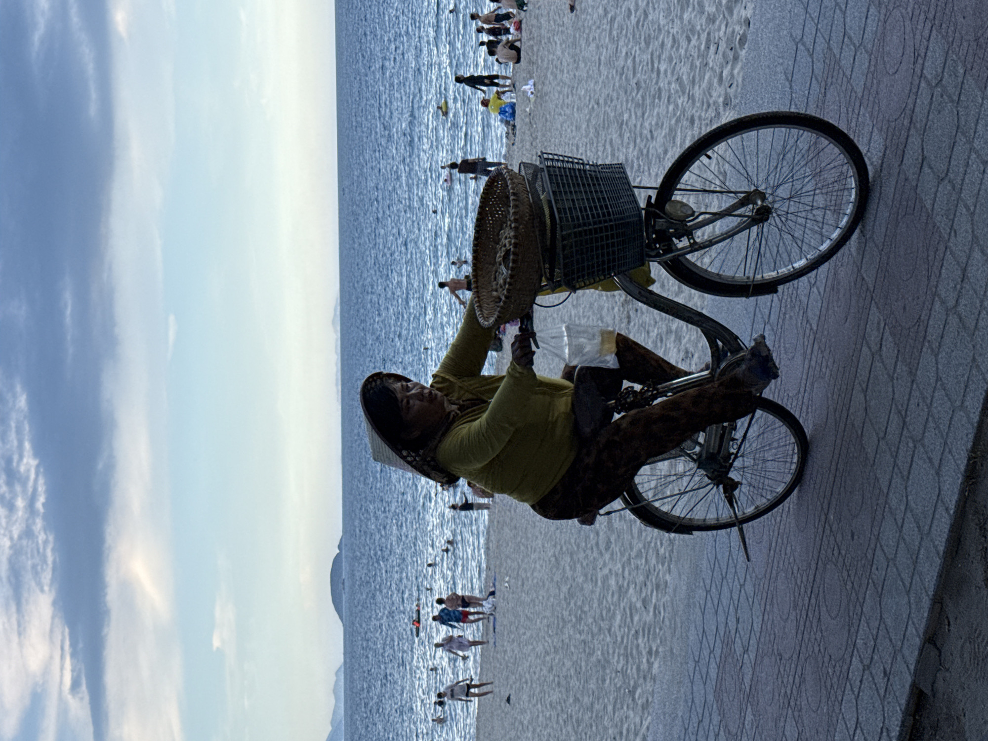 This image depicts a beach scene in Nha Trang, Vietnam. The main subject is a woman wearing a conical hat and a yellow long-sleeved shirt, along with patterned pants, pushing a bicycle. The bicycle has a large basket attached to the front, which appears to hold a woven item, possibly a hat or basket, and some plastic bags. The woman is positioned on a paved walkway that runs alongside the sandy beach and the sea.
In the background, the ocean is visible, with gentle waves and a hazy horizon. A number of people are scattered on the beach and in the water, suggesting a leisurely day. Some are standing, while others appear to be swimming or wading. The sky is mostly clear with some soft clouds, indicating fair weather. The time of day appears to be late afternoon or early evening, given the soft lighting. The overall mood of the scene is relaxed and typical of a coastal tourist destination. There is no visible text in the image.