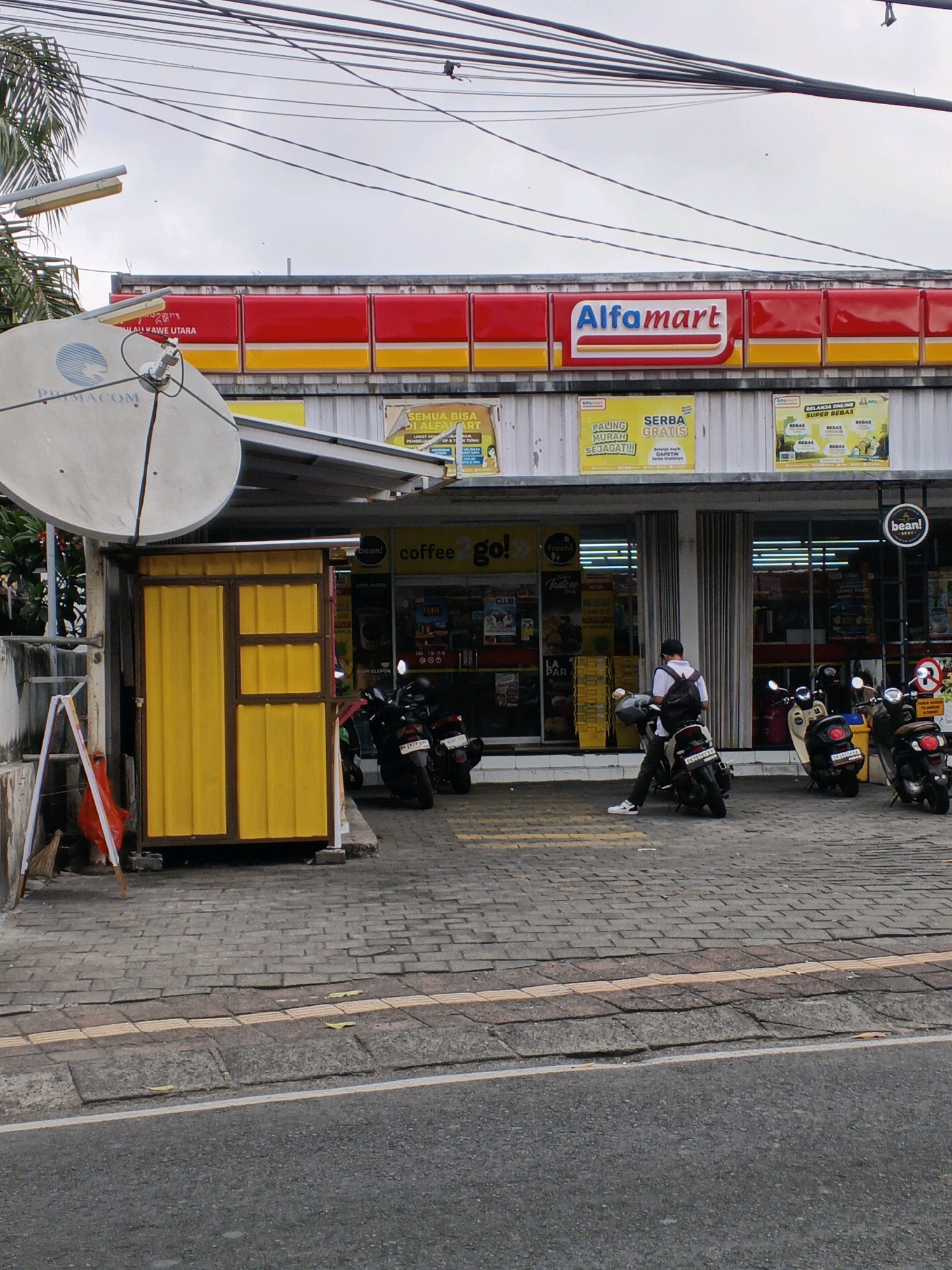 The image depicts an Alfamart convenience store located in Denpasar, Indonesia. A satellite dish, possibly for cable television, is mounted on the left side of the frame. In front of the store, a small yellow booth is visible. Several motorcycles are parked outside the store, and a person is seated on one of them, facing away from the camera, wearing a backpack. The ground is paved with interlocking bricks, and a road runs in front of the sidewalk. The sky is overcast. Various advertisements are displayed on the store's facade.