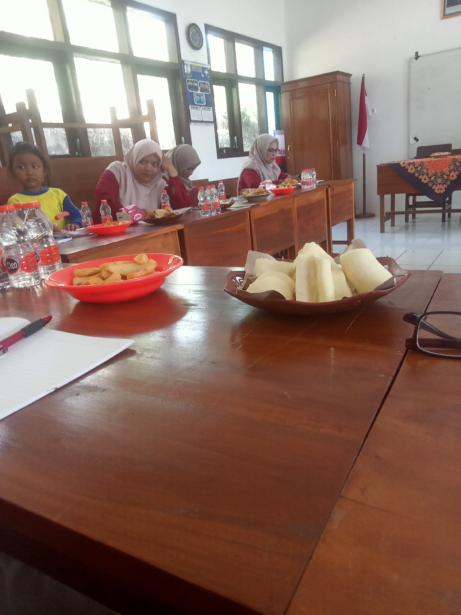 The image shows a meeting or gathering in a room in Pakel, Indonesia. There are four individuals visible: a young girl in a yellow shirt, and three women wearing hijabs and modest clothing. The women appear to be seated at a wooden table, engaged in conversation or observation. On the table, there are several bottles of "Cleo" bottled water, some red bowls containing snacks, and a bowl filled with pieces of what appears to be boiled cassava or a similar root vegetable. A notebook and a pen are also on the table, suggesting note-taking may have occurred. In the background, there is a large wooden cabinet, a whiteboard, and a Indonesian flag. The room has large windows letting in natural light, and the furniture is predominantly made of wood. The overall atmosphere suggests a formal or semi-formal gathering in an educational or community setting.