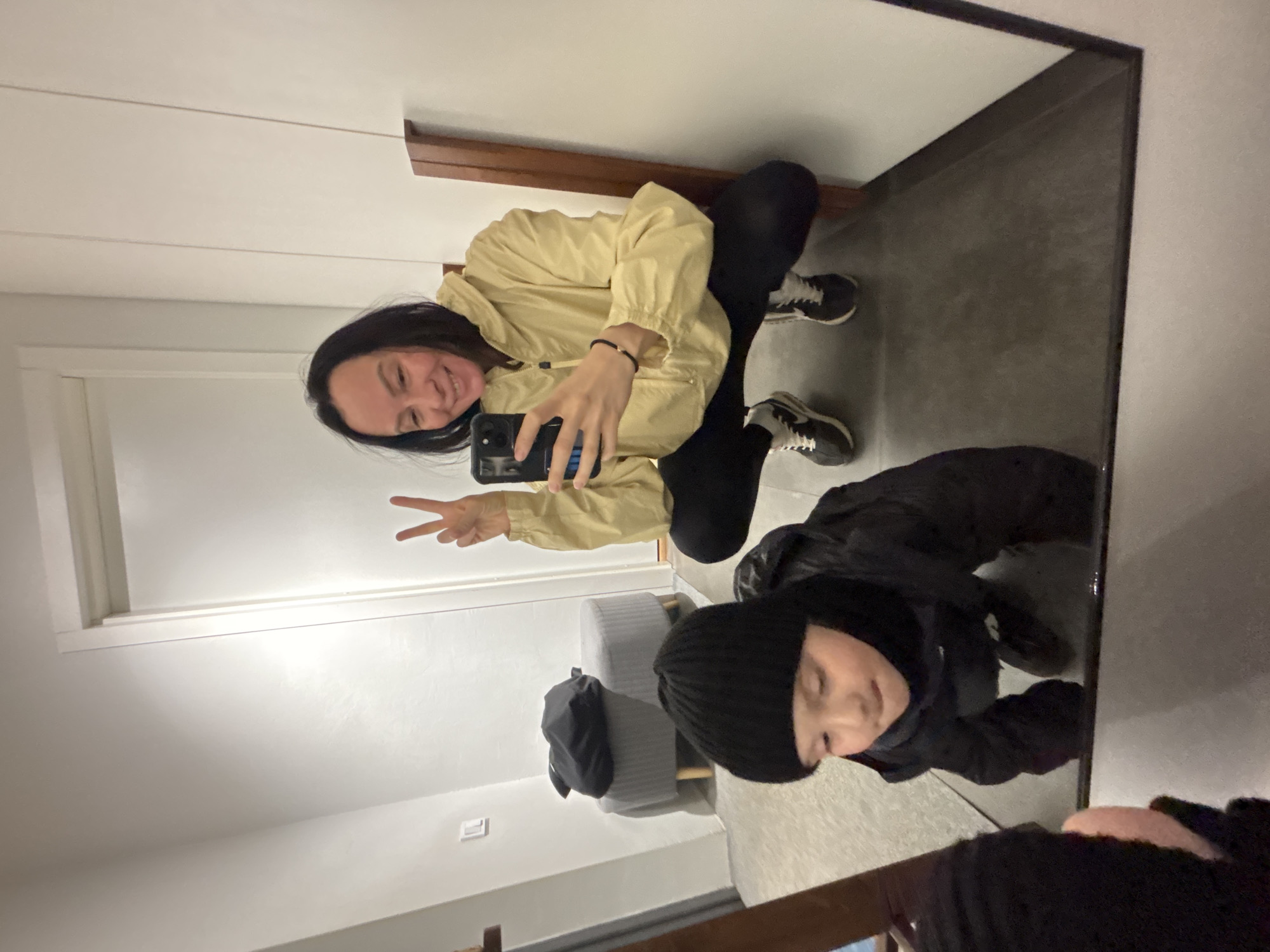A full shot depicts a woman taking a selfie with her reflection in a mirror. The woman is squatting, wearing a yellow jacket and black pants, and holding up two fingers in a peace sign. In the mirror's reflection, another person is kneeling, wearing a black jacket and beanie. The background includes a gray ottoman with a black bag, white walls, and gray flooring. The specific location in Chaikovsky, Russia cannot be determined from this image.