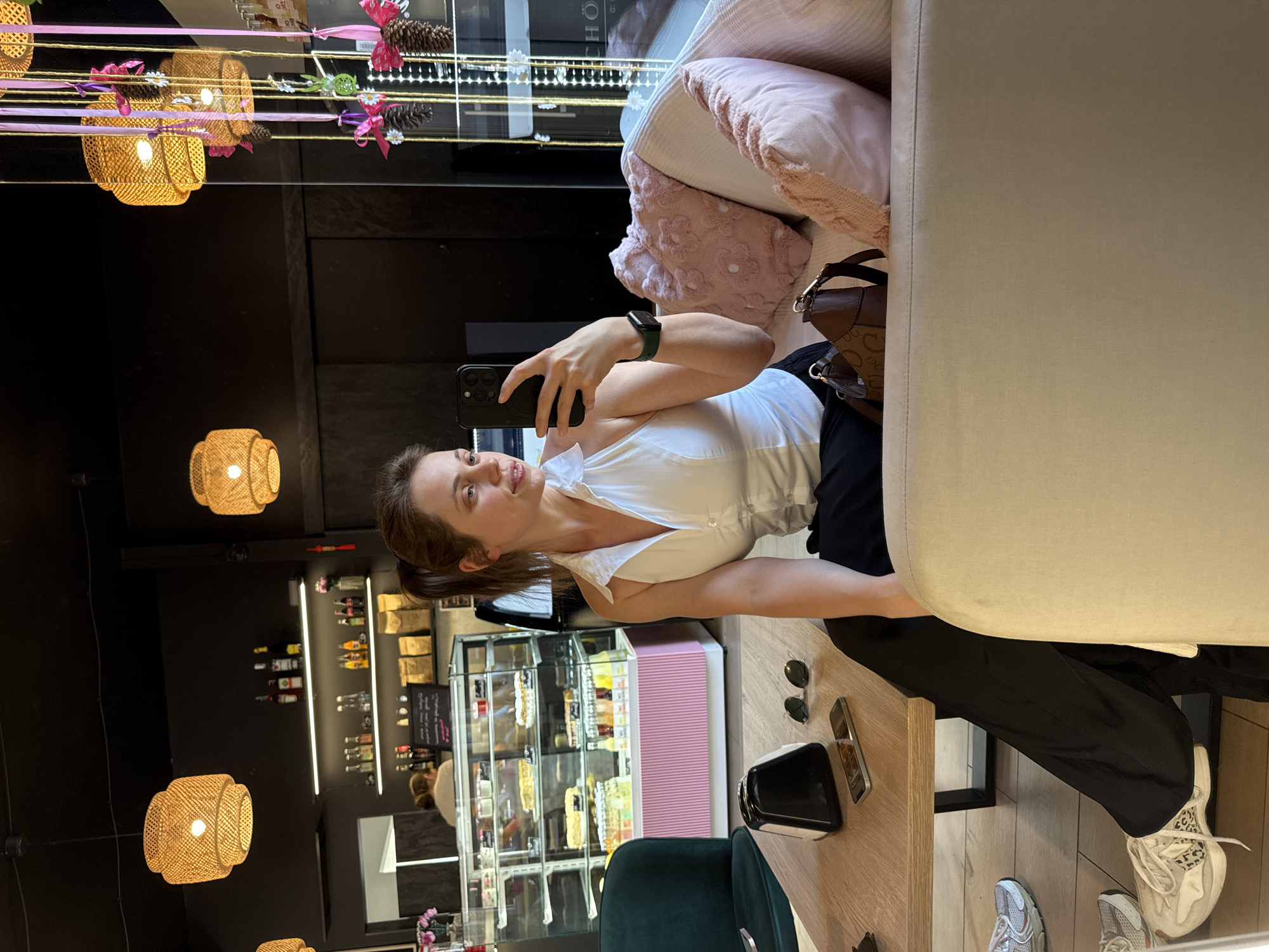 A young woman is taking a selfie in a cafe. She is wearing a white V-neck top and black pants, and she has her hair tied back in a ponytail. She is holding up her phone to take the photo, and she is smiling. The cafe is decorated with woven lamps and other rustic elements. In the background, there is a display case filled with pastries and other food items. The woman is sitting on a couch, and there is a handbag next to her. On the table in front of her, there are sunglasses and a phone. The image appears to have been taken during the day. The overall mood of the image is casual and relaxed.