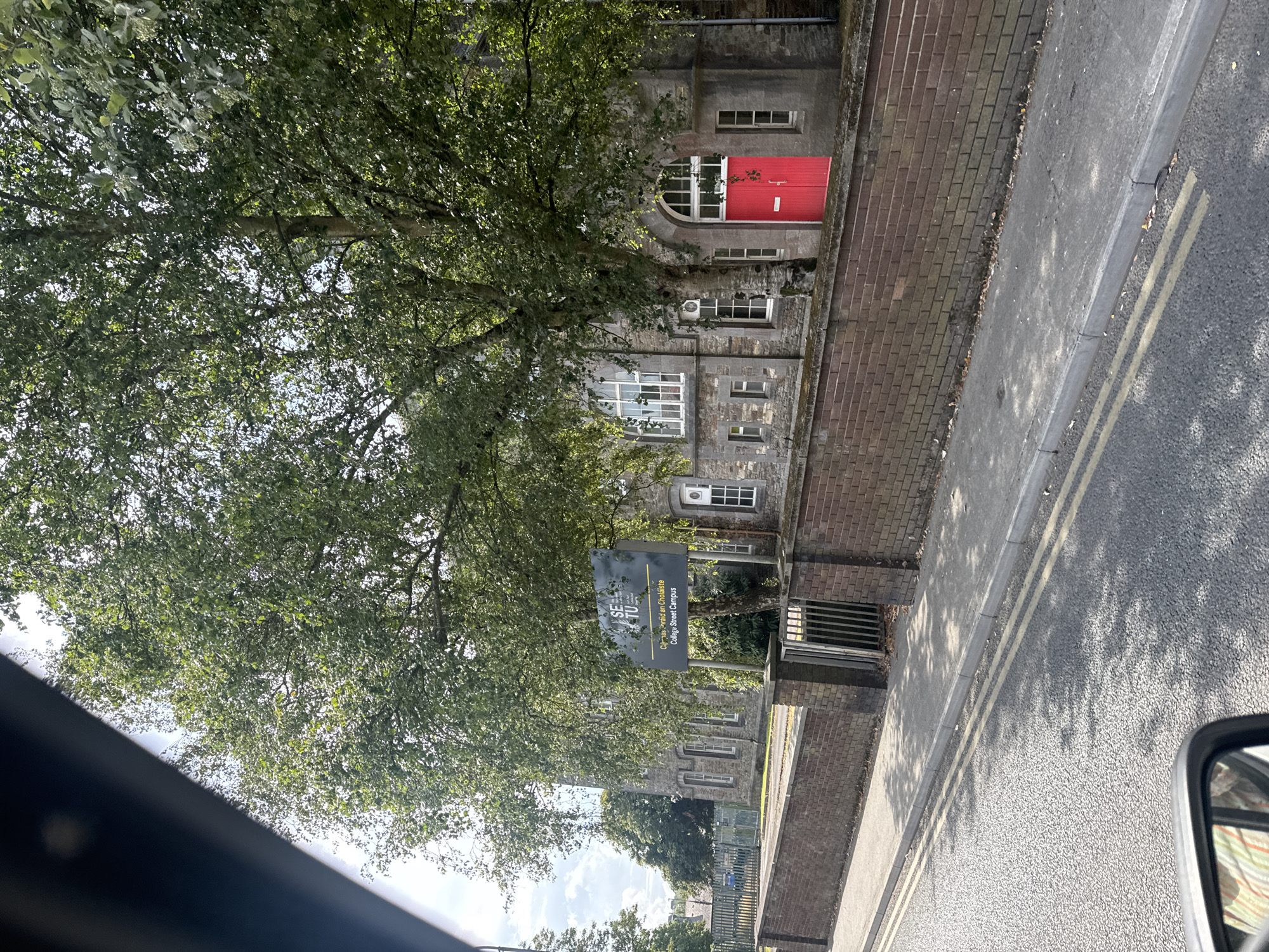 The image shows the "SETU" sign for the College Street Campus of the South East Technological University in Waterford, Ireland. The sign is dark gray with white lettering and has the text "Caritas an Choláiste" and "College Street Campus" below the main logo. A stone building with several windows and a bright red door is visible behind the sign and a low brick wall. The building appears to be an older structure, possibly an educational institution. Trees with green foliage are abundant, casting shadows on the sidewalk and road. The road in the foreground has double yellow lines and a sidewalk runs along the brick wall. The image appears to be taken from a vehicle, as a portion of a side mirror is visible in the bottom left corner.