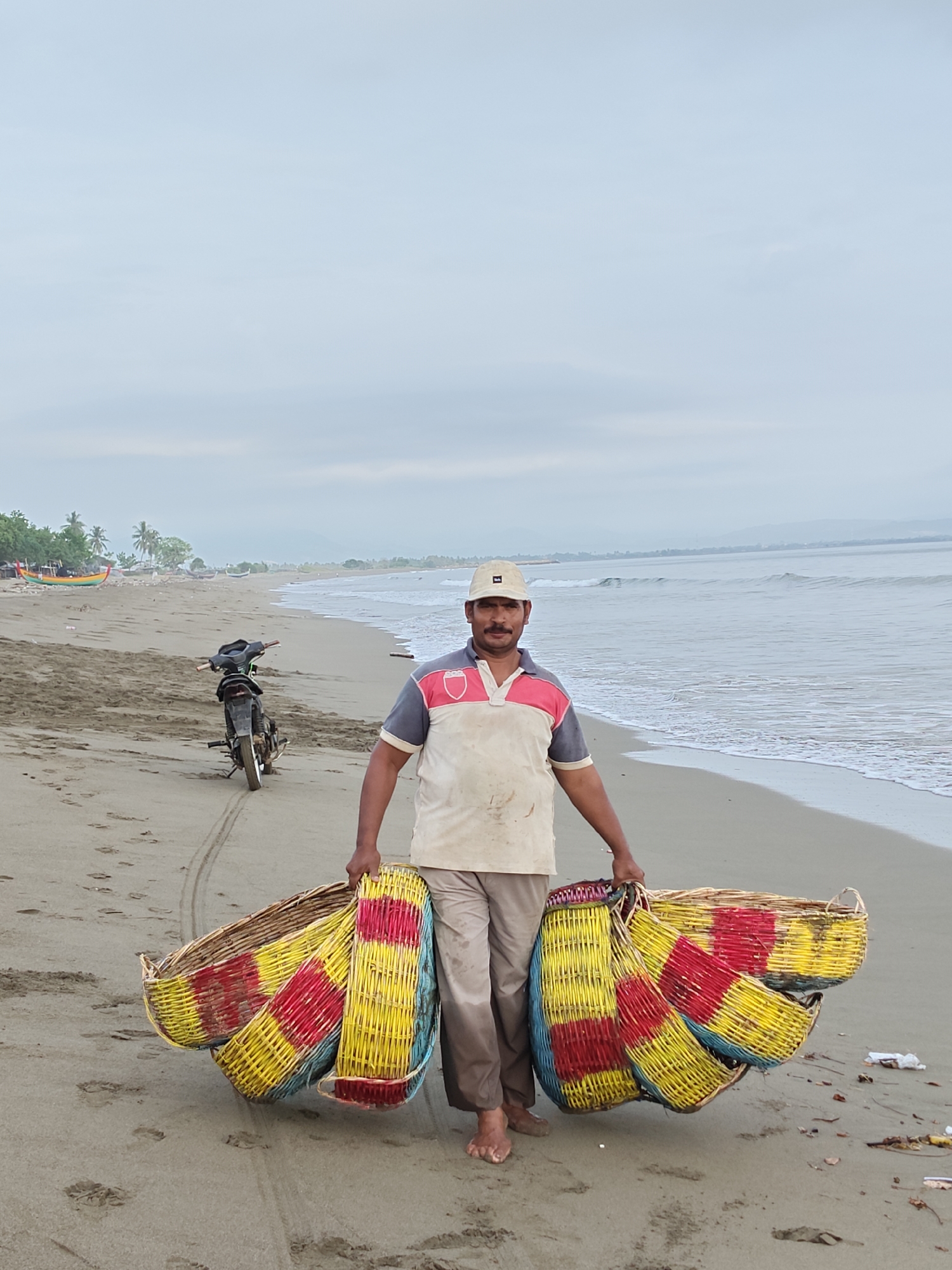 A man is walking on a beach in Curehtunong, Indonesia, carrying several woven baskets. He is wearing a light-colored polo shirt with red and grey accents and a light-colored baseball cap. His pants are a neutral tan color, and he is barefoot. He is holding multiple, colorful woven baskets, predominantly yellow and red, in front of him. Behind him, a motorcycle is parked on the sand. The ocean is visible to his right, with gentle waves rolling onto the shore. The beach is sandy with some debris scattered on it. In the distance, there are trees and a hazy mountainous landscape under a pale, overcast sky.
